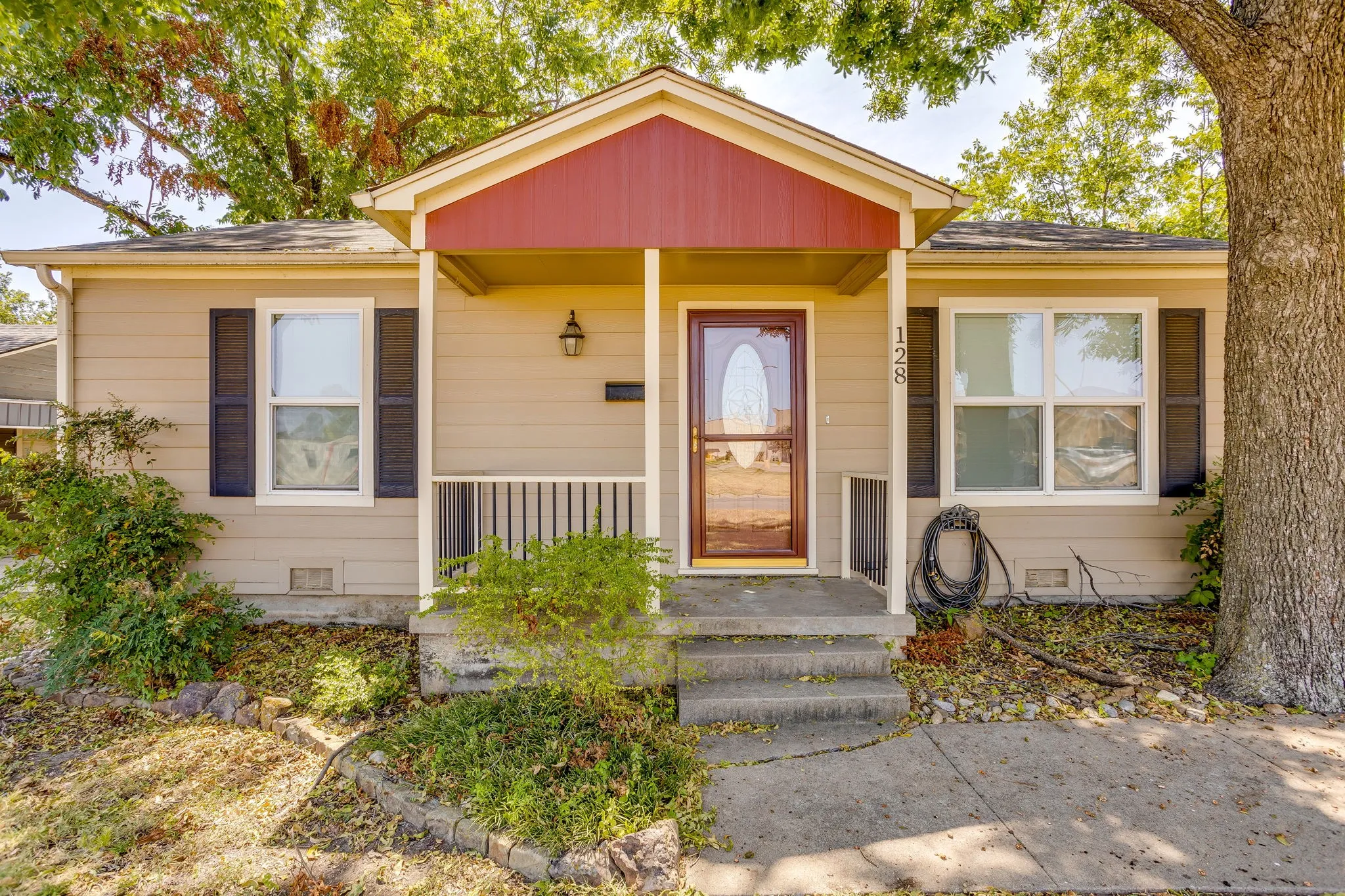 Front exterior featuring light-colored siding, a covered porch with a dark red accent, a paneled wood-finish entry door with a storm door, and dark shutters on multiple windows