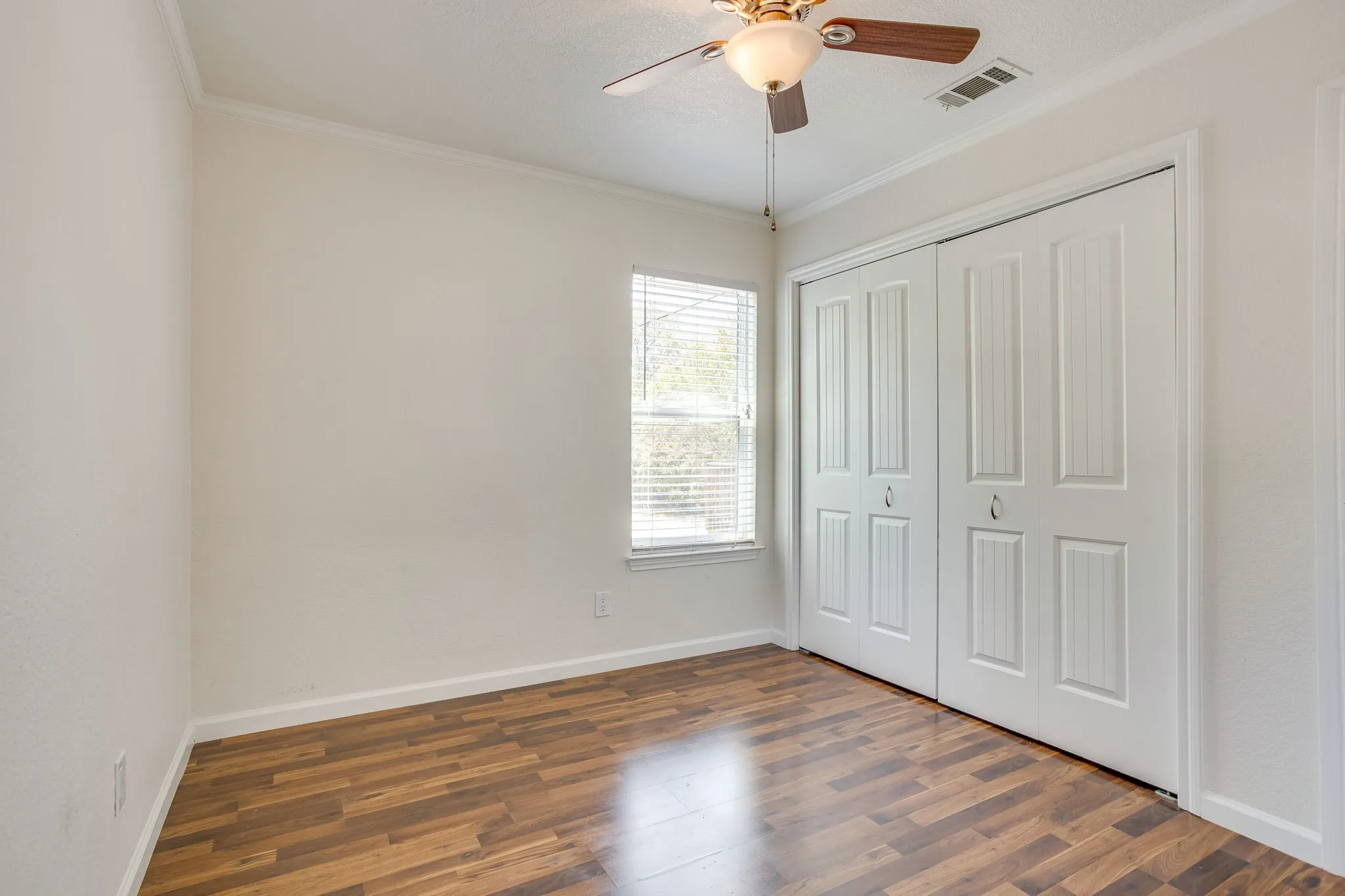 Wood laminate flooring, white walls, double bi-fold closet doors, crown molding, and a ceiling fan with integrated lighting