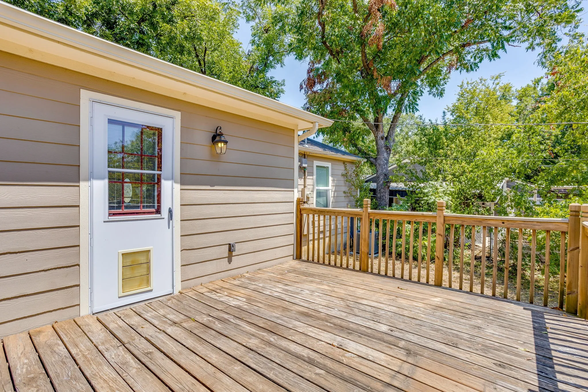 Rear exterior deck with wood flooring and railing, featuring a mounted outdoor light fixture and a white door with glass panels