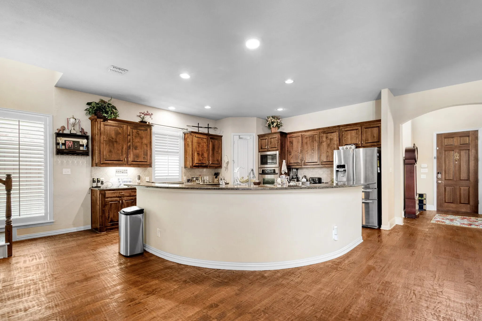 Expansive wood-finish flooring, recessed lighting, wood cabinetry, built-in stainless steel appliances, and a curved breakfast bar with a stone countertop