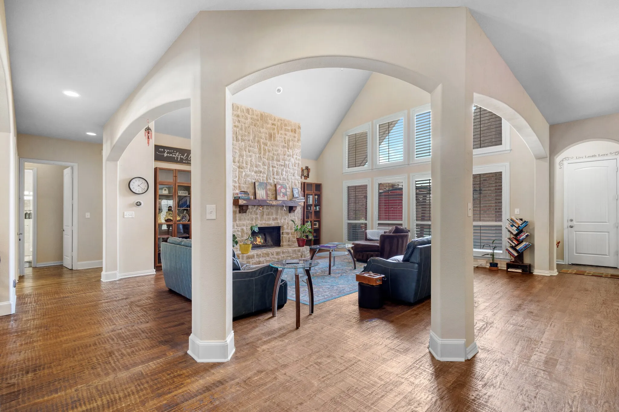 Vaulted ceiling living space featuring a stacked stone fireplace, large windows with plantation shutters, arched doorways, and wood-finish flooring