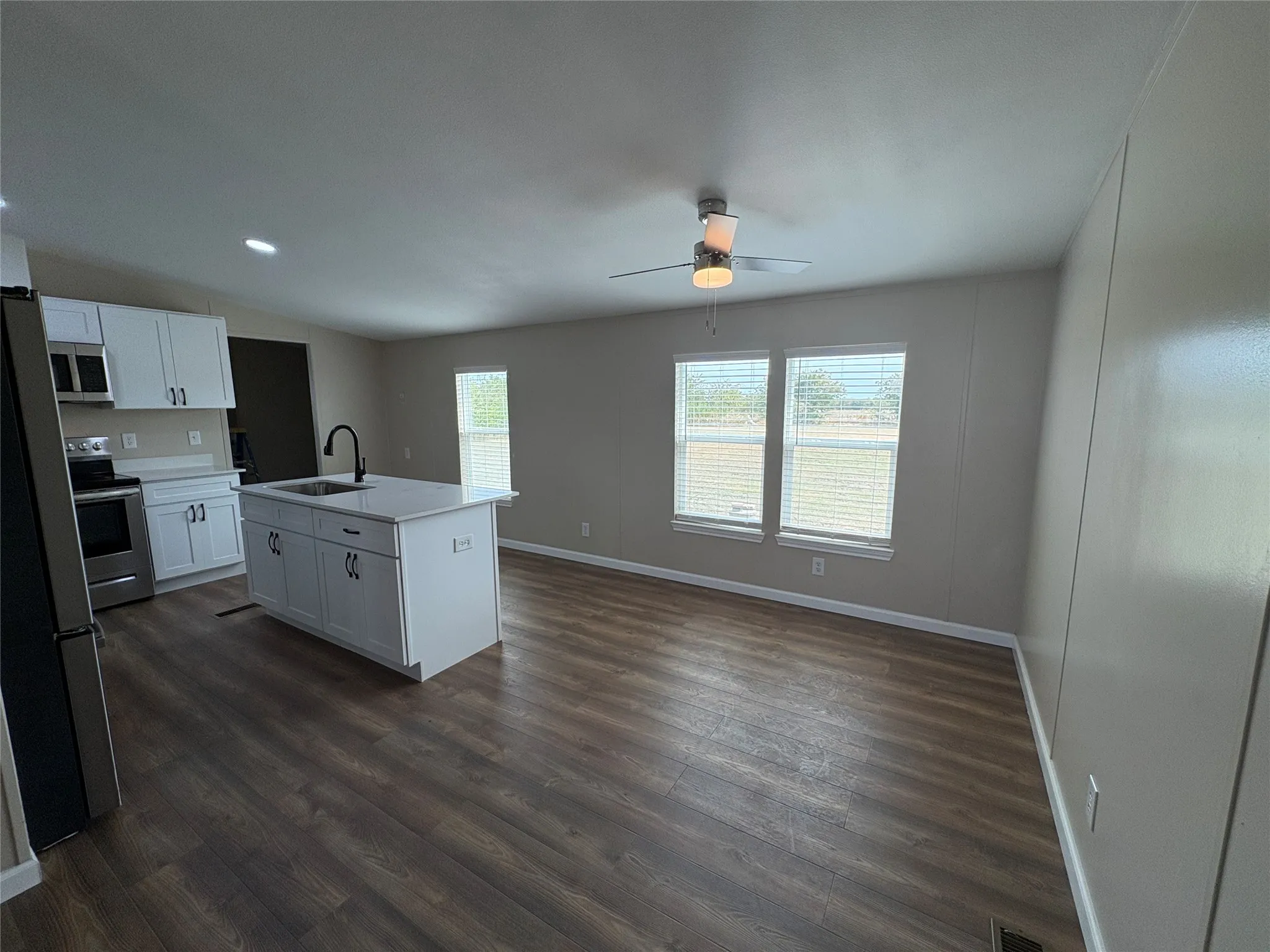 Open-concept living space with wood-finish flooring, a kitchen island with integrated sink, white cabinetry with dark hardware, stainless steel appliances, and a ceiling fan