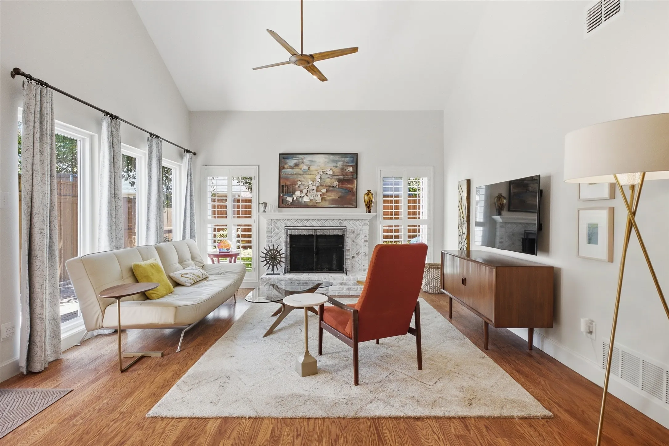 The living room earns its place at the heart of the home — vaulted ceilings, plantation shutters, hardwood floors, and a white herringbone brick fireplace with a classic mantel. Light and airy in every season.
