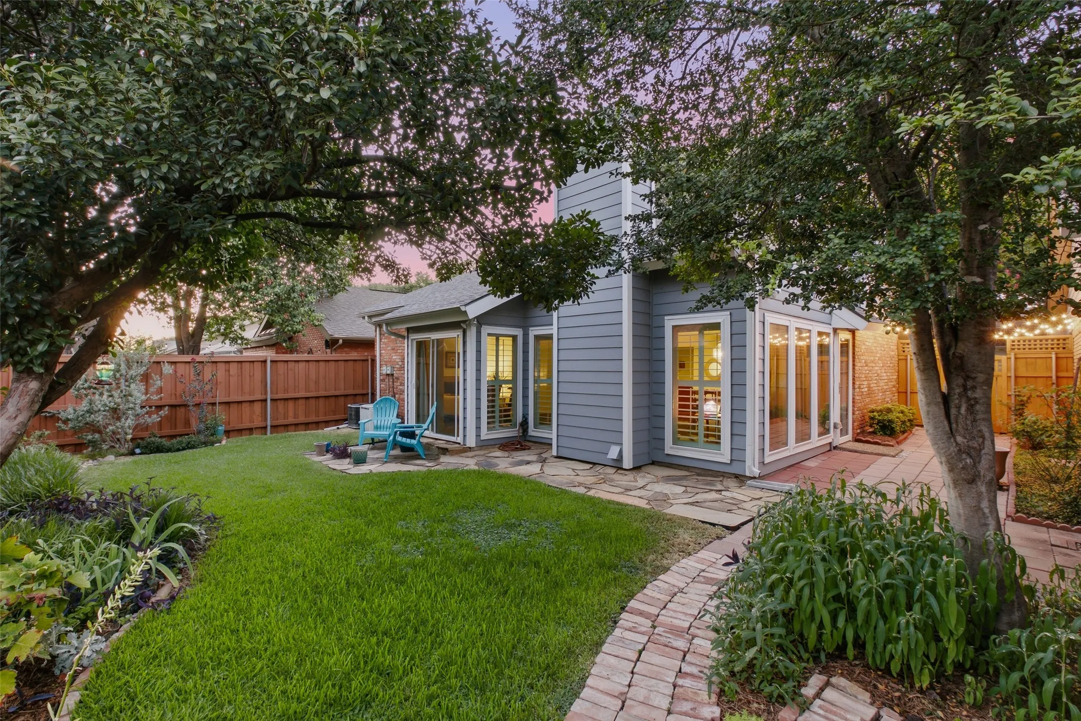 The backyard from across the lawn — well-maintained, fully fenced, with established landscaping along the perimeter and a warm glow from the home's rear windows.