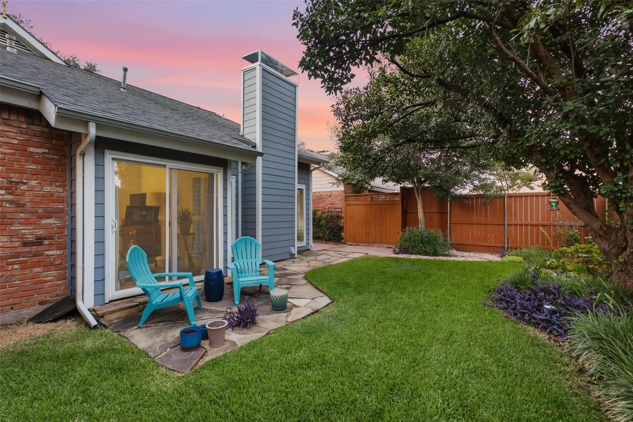 The backyard patio at dusk — flagstone pavers, a private fenced yard, mature shade tree, and colorful garden beds. A quiet spot that gets better as the sun goes down.