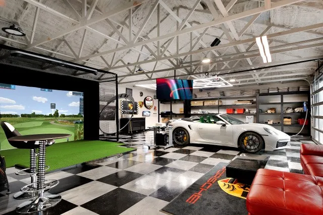Spacious garage with an exposed truss ceiling, checkered flooring, full-height built-in shelving, a golf simulator, and a full-view garage door