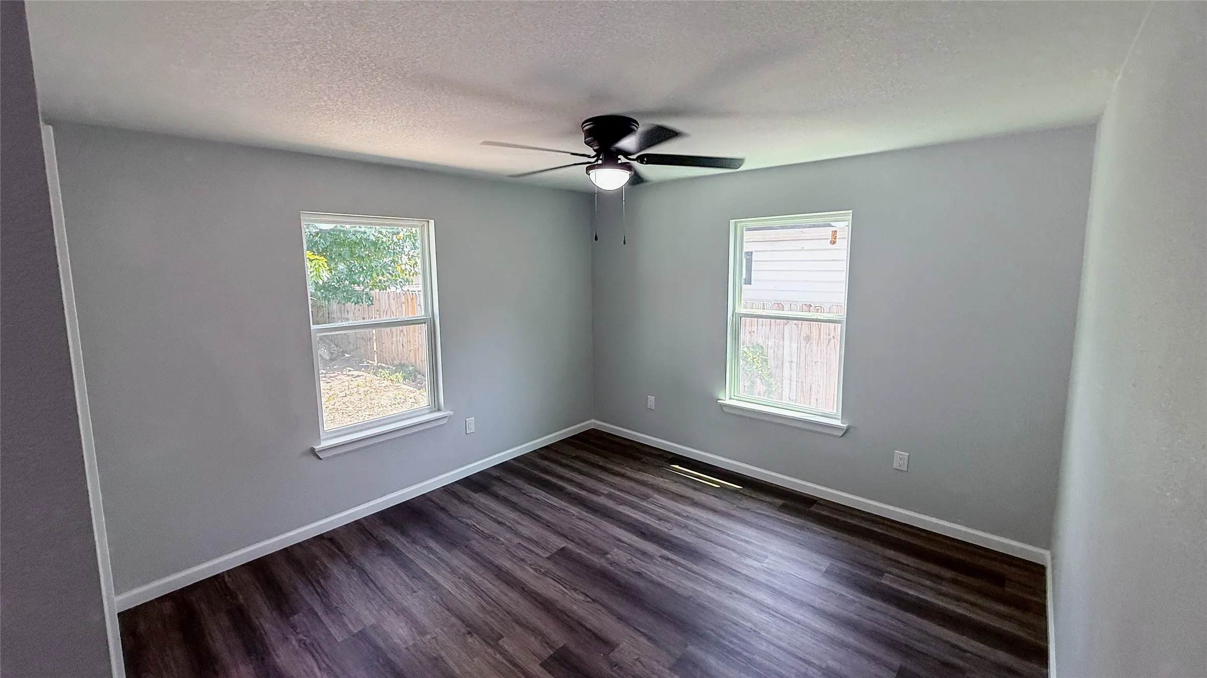 Wood-finish flooring, light gray wall paint, two windows, and a ceiling fan with integrated lighting