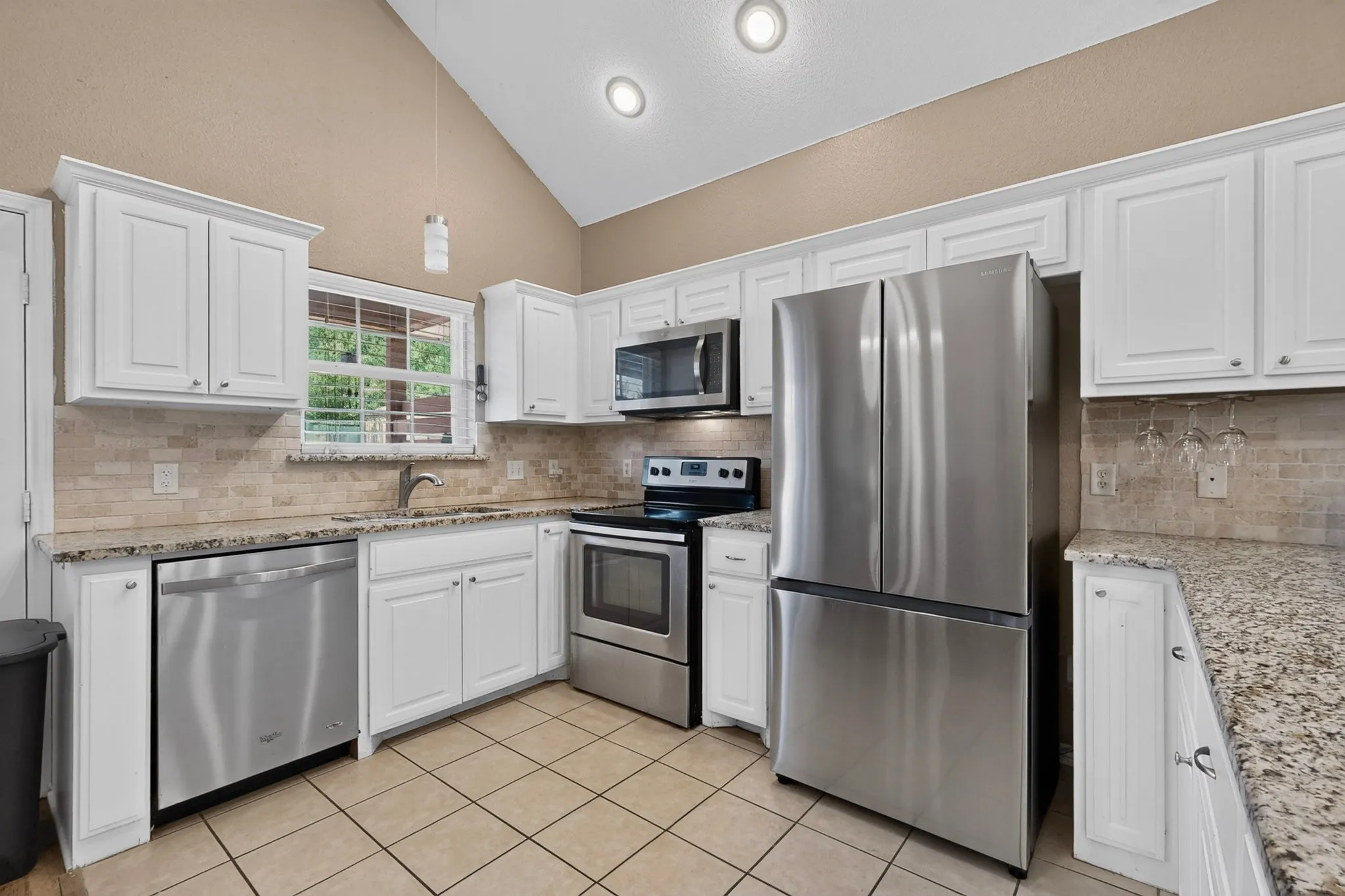 Vaulted ceiling kitchen featuring white cabinetry, light-toned tile flooring, light-toned backsplash, and stainless steel appliances
