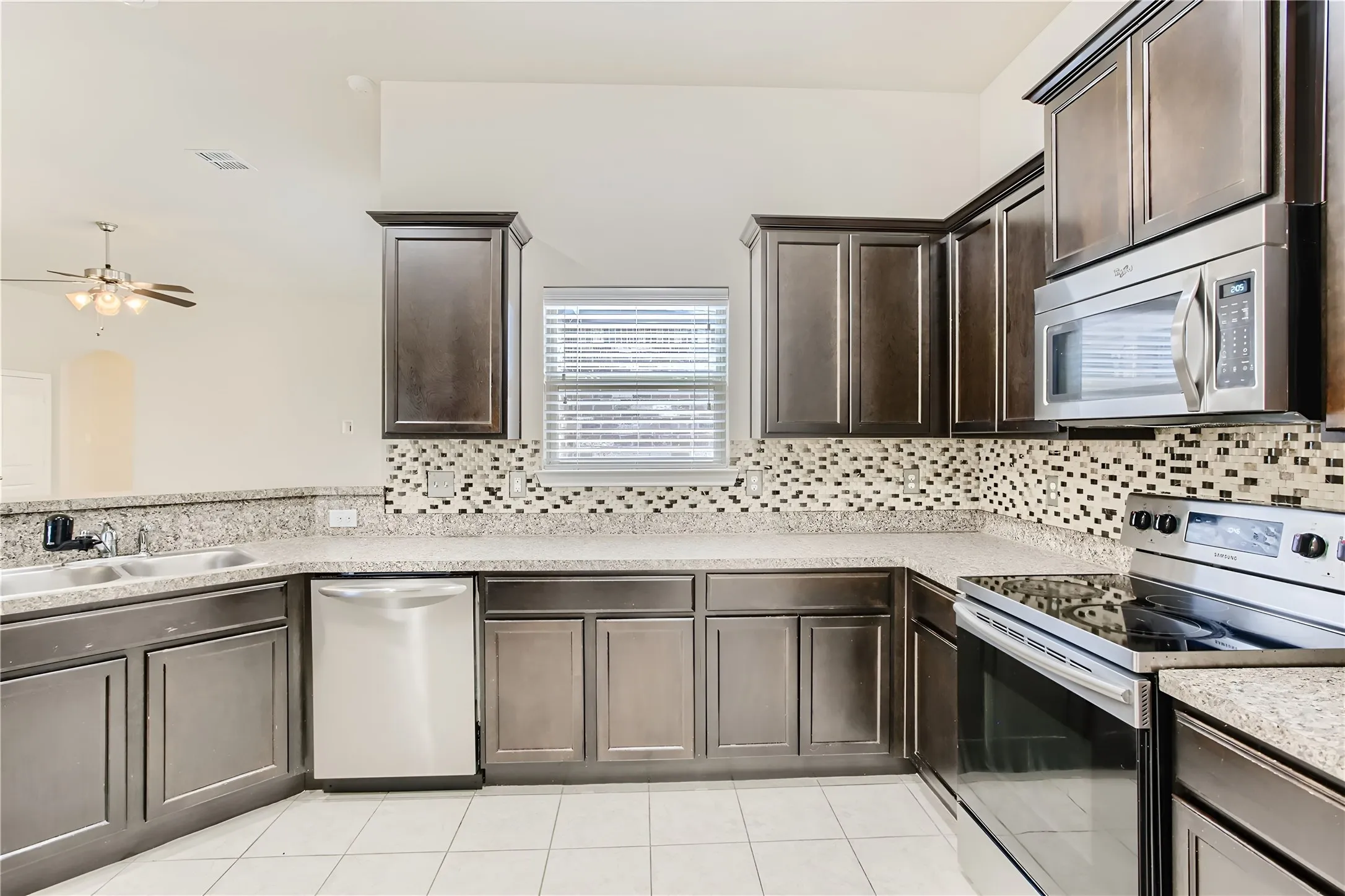 Kitchen featuring dark wood-finish cabinetry, light-toned granite-style countertops, stainless steel appliances, a mosaic tile backsplash, and white tile flooring