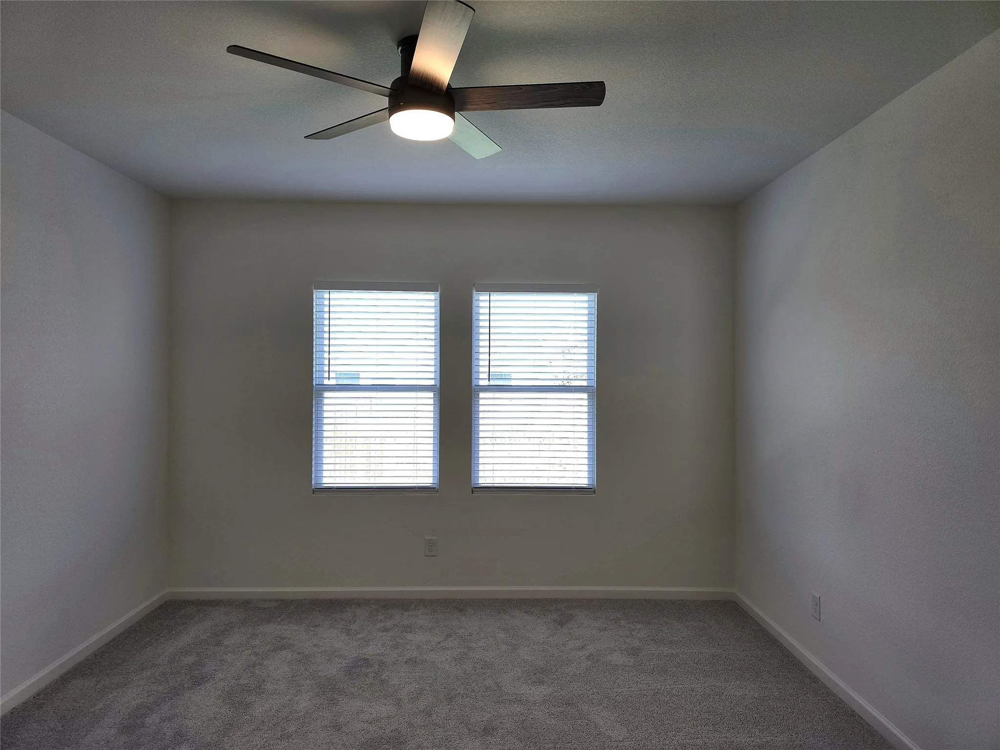 Neutral wall color, two windows with blinds, ceiling fan with integrated lighting, and carpet flooring