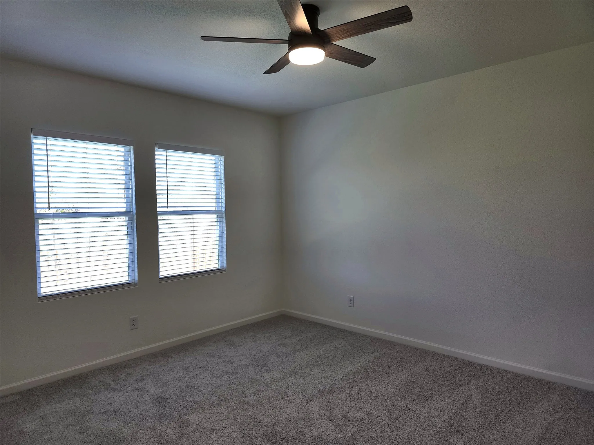 Two windows with horizontal blinds, overhead ceiling fan with integrated lighting, and gray carpeted flooring