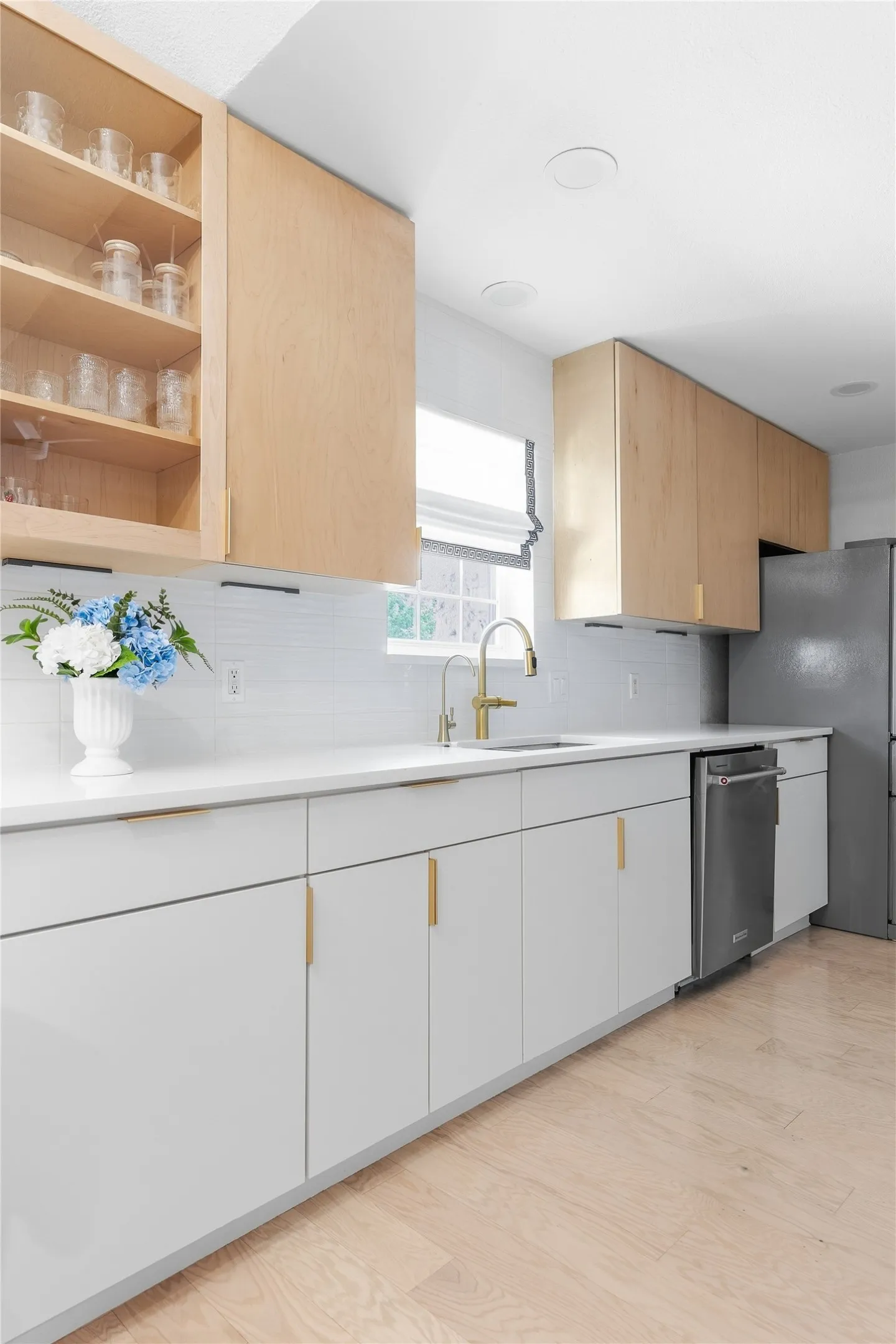 Modern kitchen featuring light wood-finish flooring, white flat-panel cabinetry, light wood-tone upper cabinetry, a white subway tile backsplash, and a gold-finish faucet