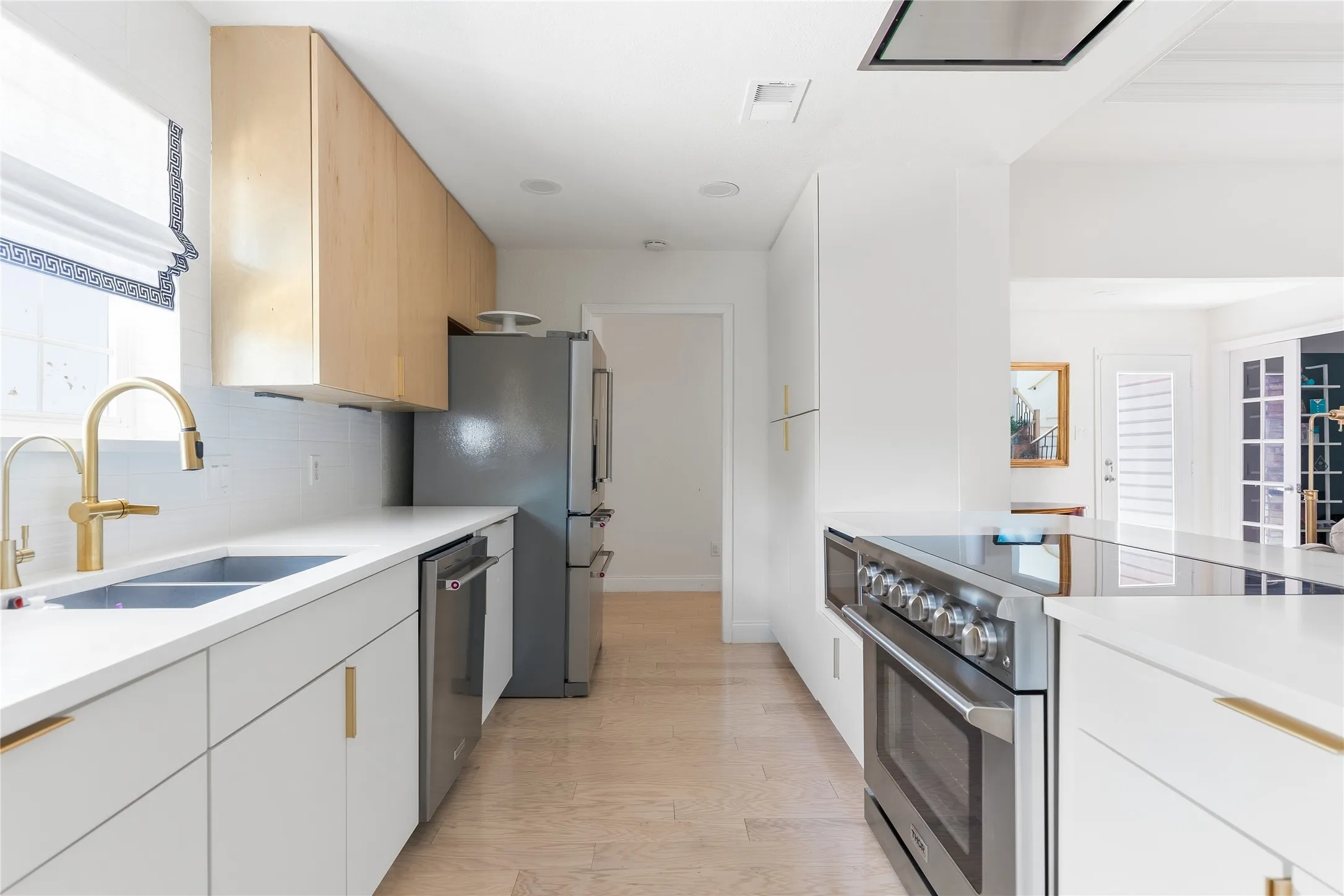 Contemporary kitchen featuring stainless steel appliances, light wood-finish flooring, white cabinetry with gold-tone hardware, and a dual-basin sink with a gold-tone faucet