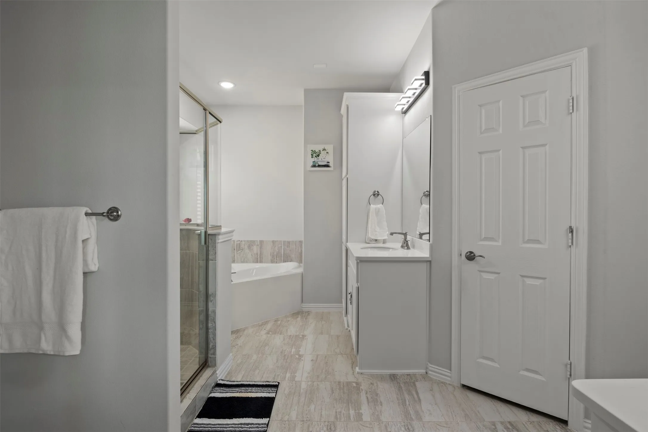 Glass-enclosed shower, built-in corner bathtub, single vanity with integrated sink, light wood-finish flooring, and recessed ceiling lighting