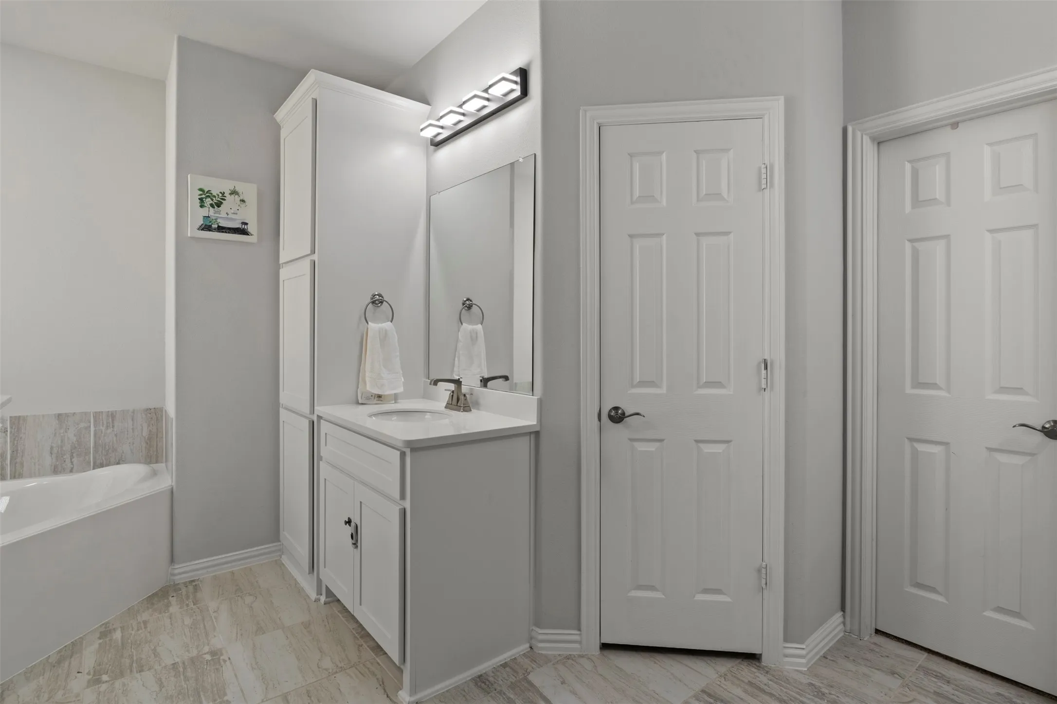 White vanity with integrated sink, modern light fixture, built-in storage cabinetry, tiled flooring, and a bathtub with tiled surround