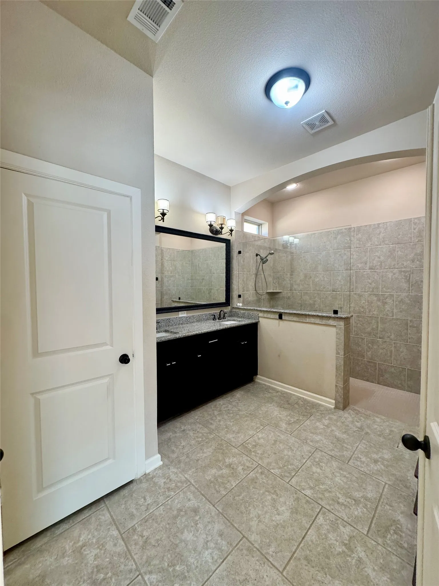 Spacious bathroom featuring large format tile flooring, a dual vanity with a dark wood-finish cabinet and light-toned countertop, and a walk-in shower with tiled walls