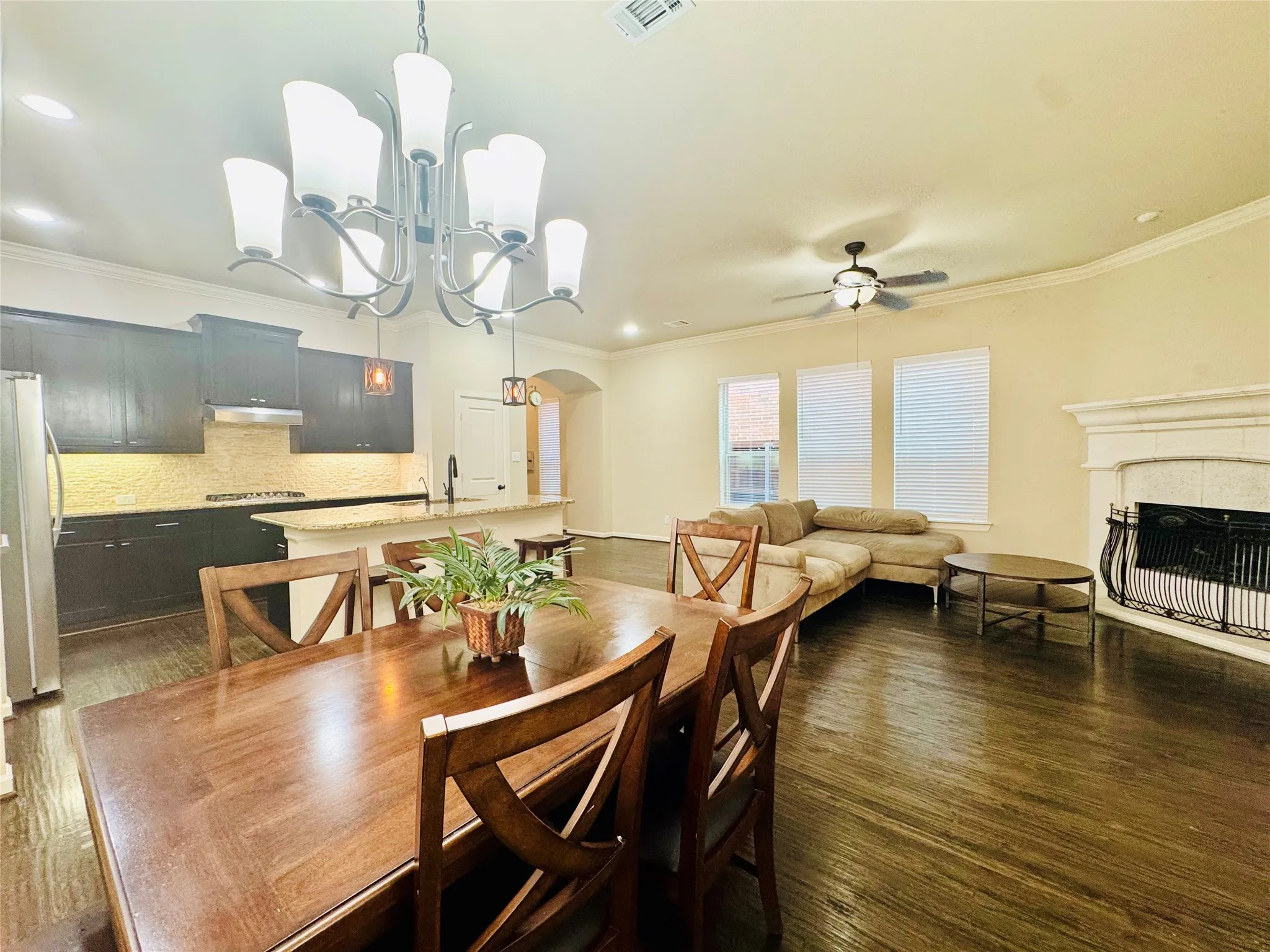 Hardwood-finish flooring throughout, kitchen with dark cabinetry, granite countertops, and tile backsplash, living area with a built-in fireplace, and a dining area with a chandelier
