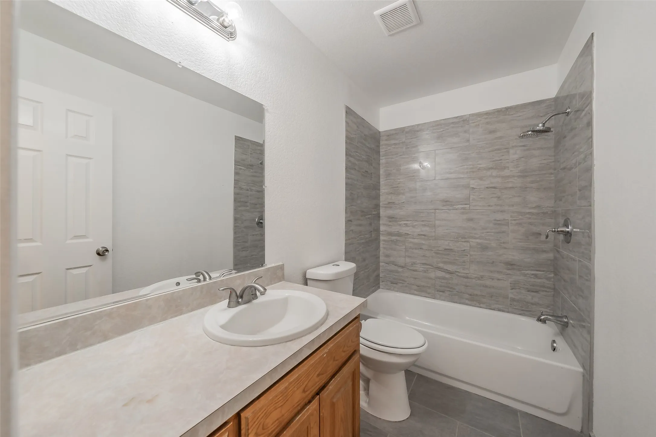 Full-width vanity mirror, wood-finish cabinetry, tile flooring, ceramic sink, and tiled shower surround with a built-in tub
