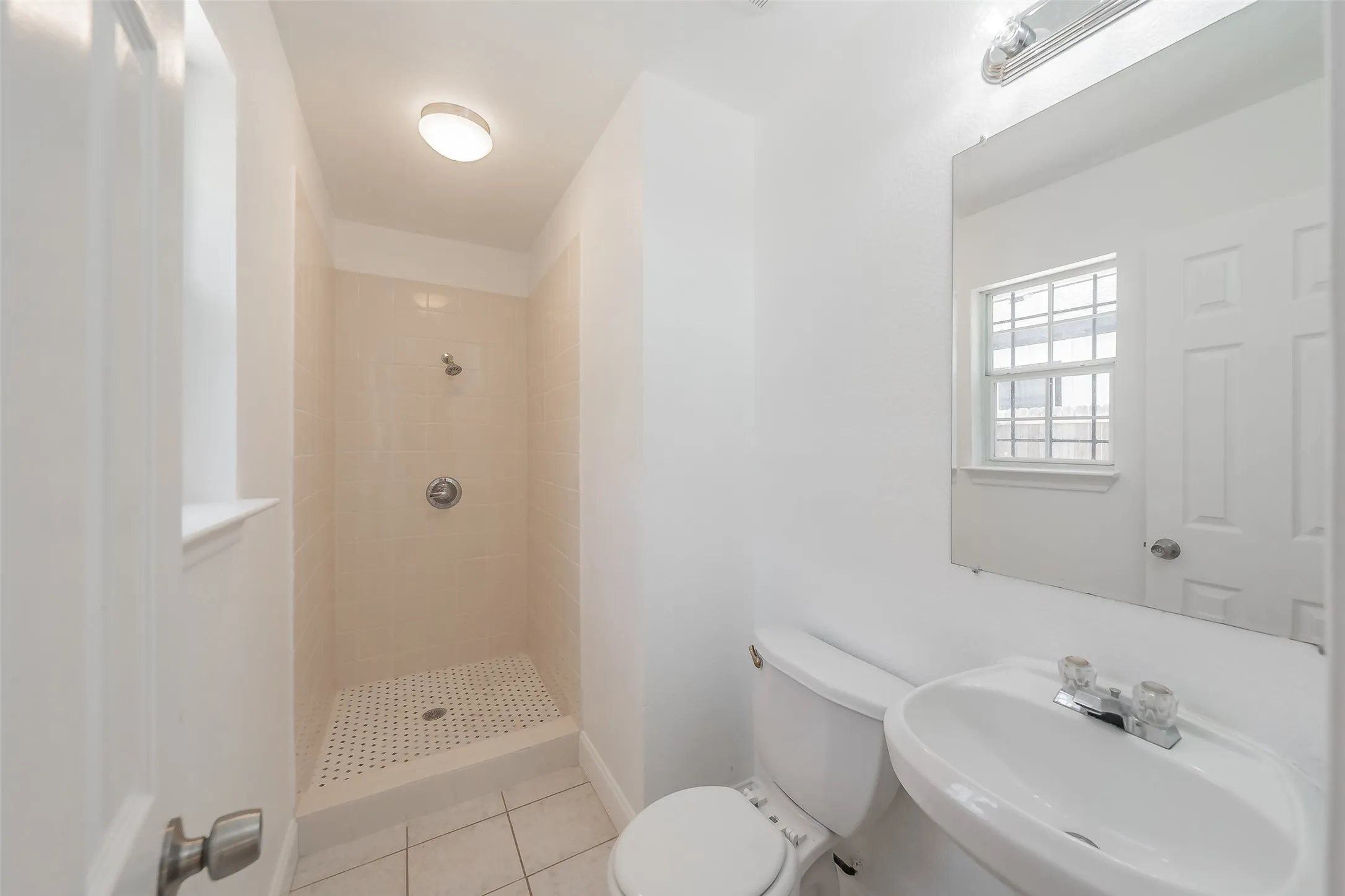 White ceramic tile flooring, walk-in shower with tiled walls, wall-mounted shower fixture, pedestal sink with chrome faucet, and a wall-mounted mirror