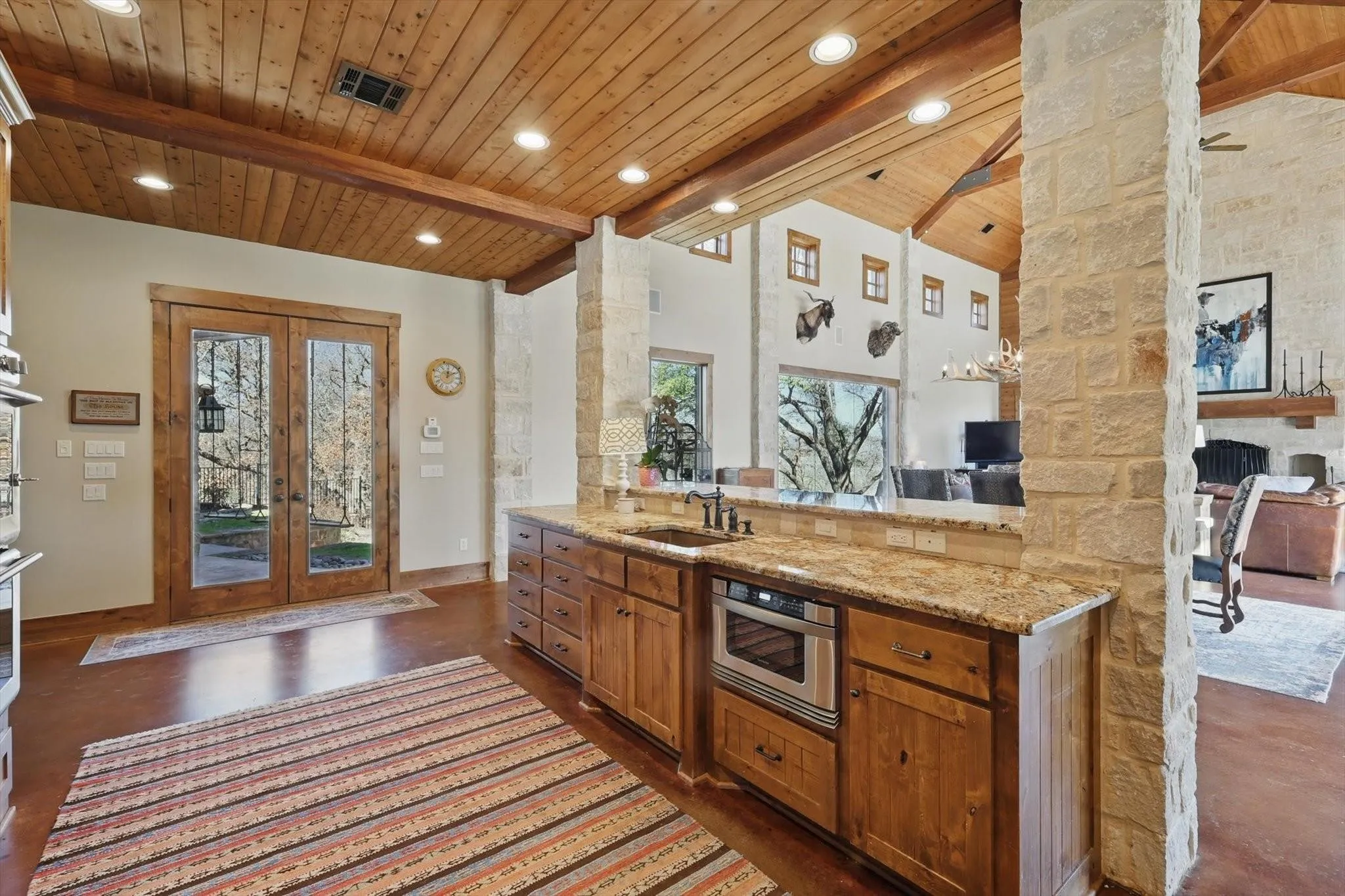 Kitchen with French doors, brown cabinetry, wood ceiling with exposed beams, light stone countertops, and open floor plan.