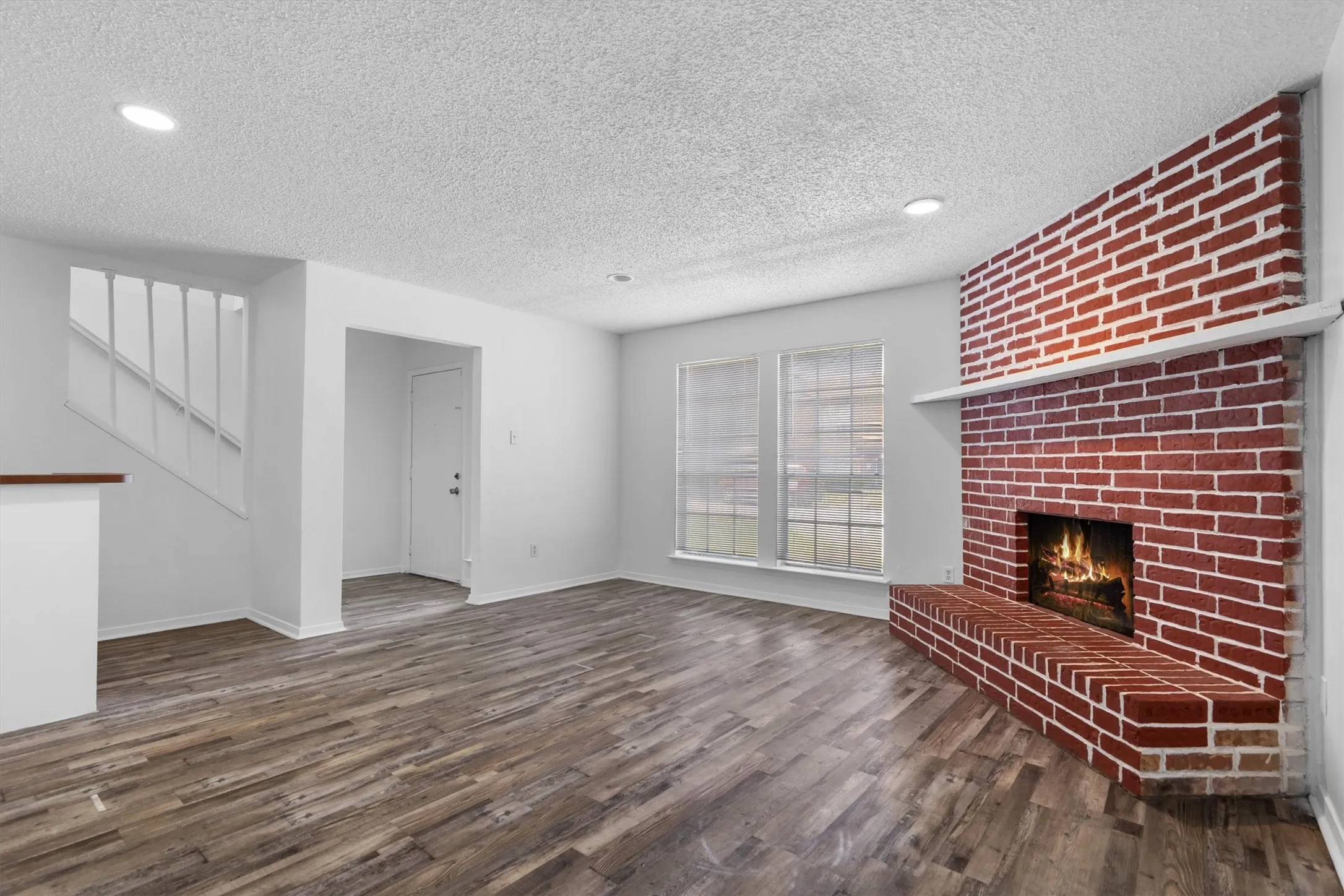 Spacious living area featuring wood-finish flooring, a red brick fireplace with a mantle, and two large windows