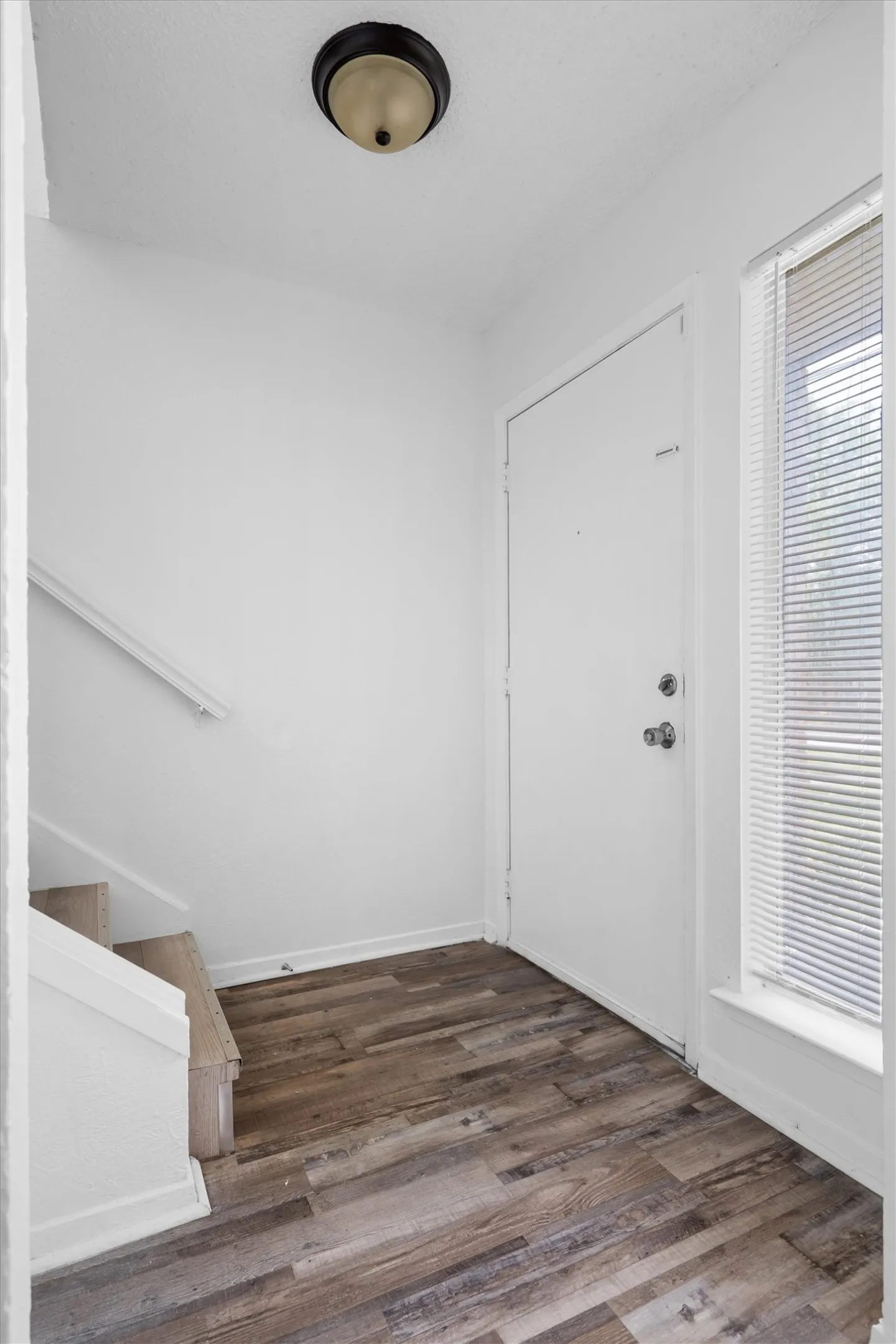 Bright entryway showcasing warm wood-finish flooring, white painted walls, a recessed ceiling light fixture, and direct access to additional living spaces