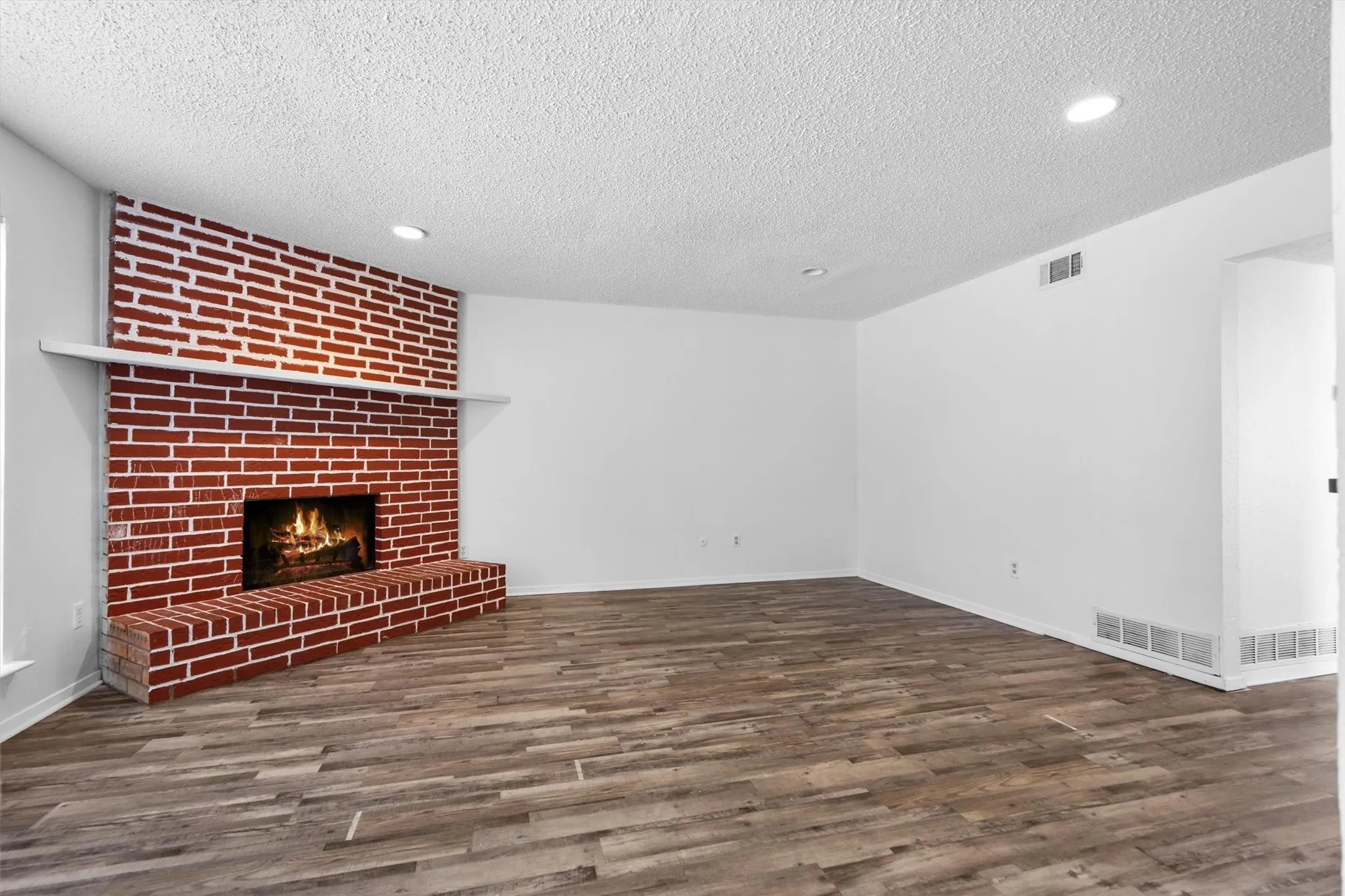 Exposed brick fireplace with a white mantel, wood-finish flooring, recessed lighting, and a textured ceiling