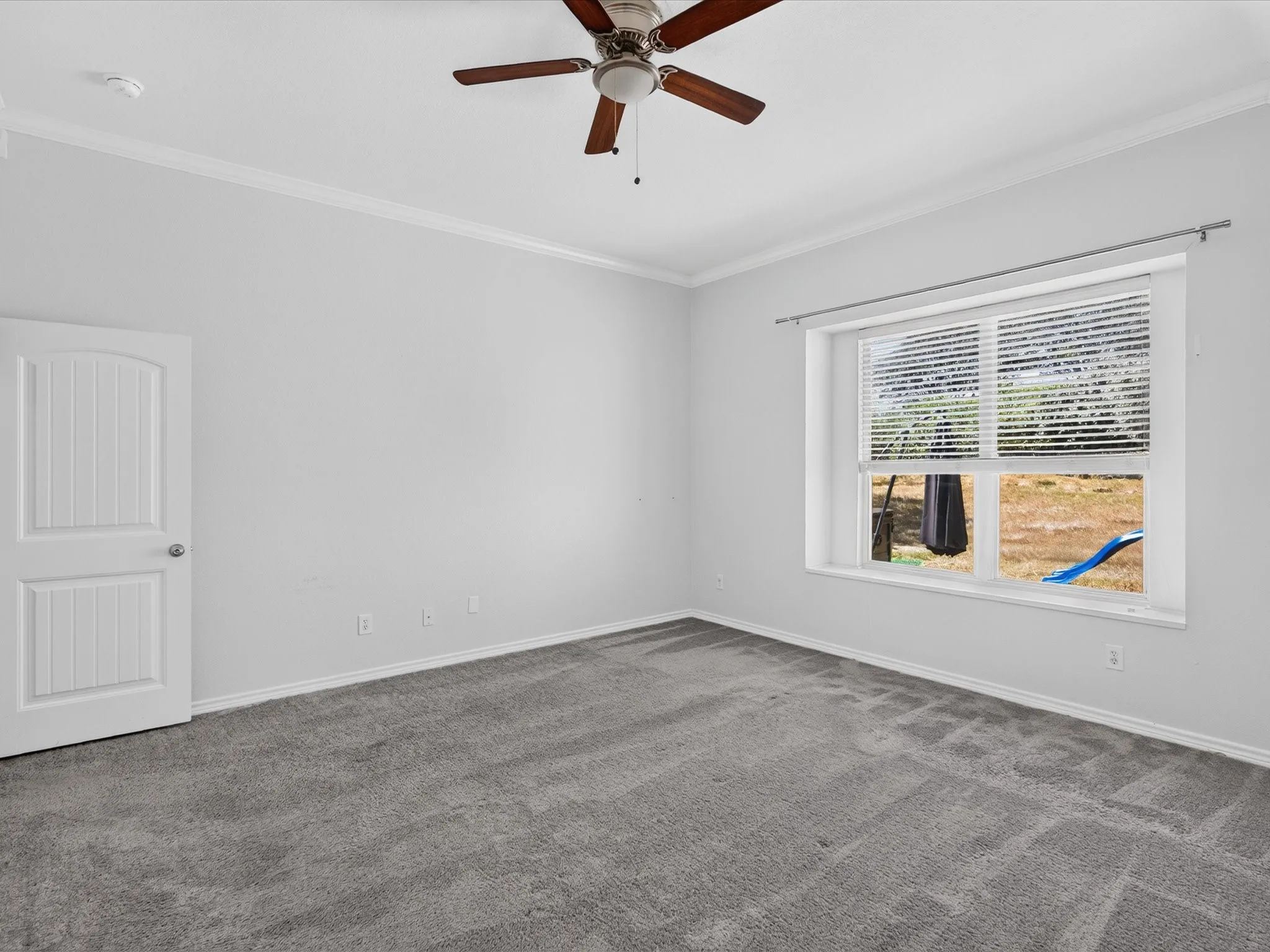 Bright and spacious bedroom featuring neutral wall finishes, plush carpeting, a ceiling fan with wood-finish blades, and large windows with plantation shutters flooding the room with natural light