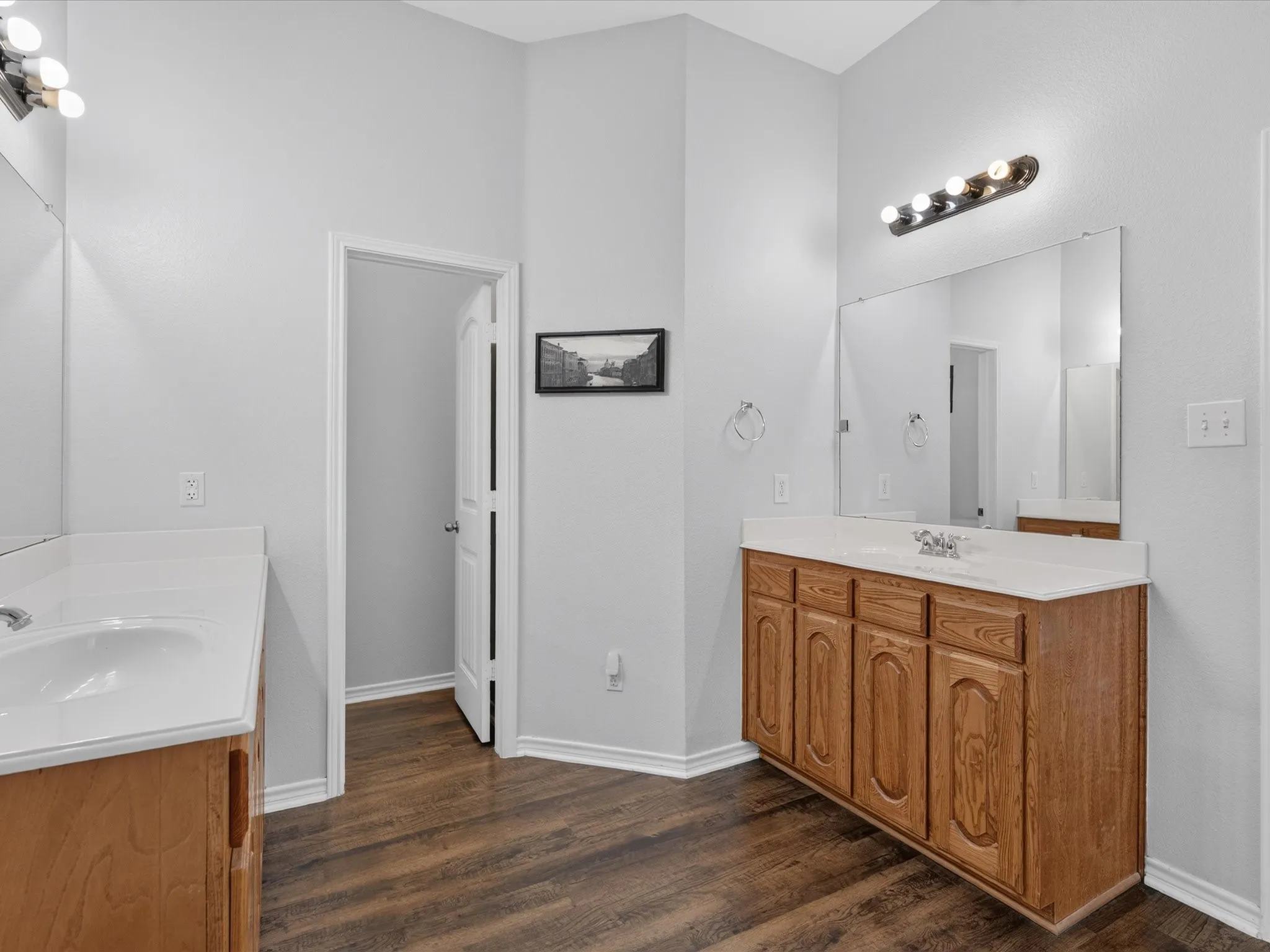 Dual vanity with white countertops, integrated sinks, wood-finish flooring, and wall-mounted lighting fixtures