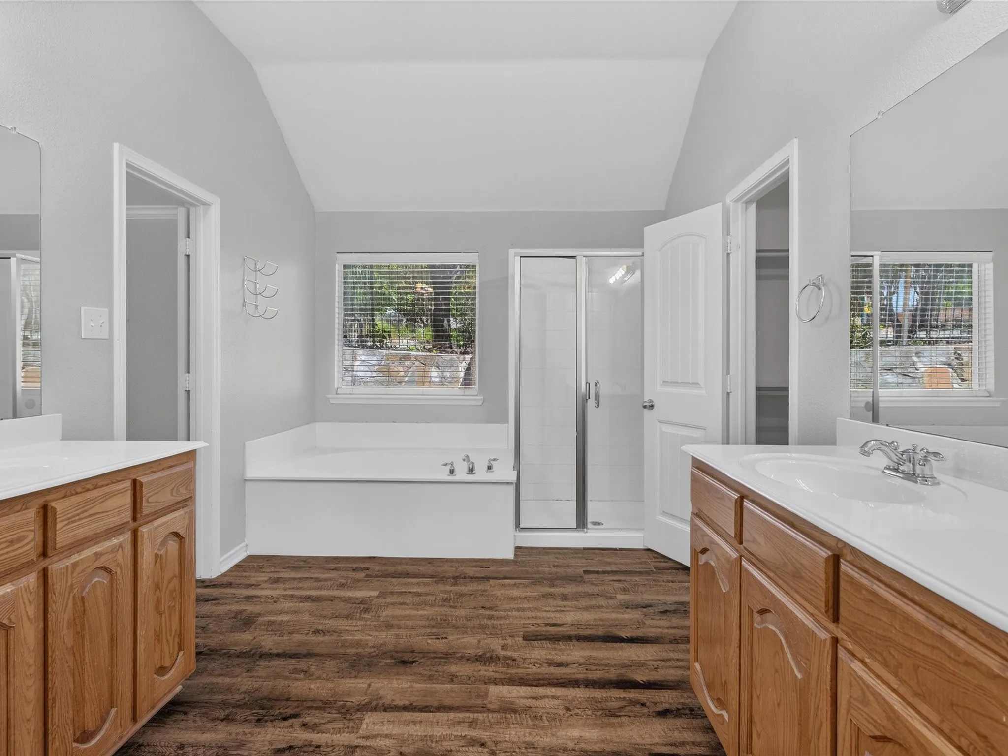 Vaulted ceiling bathroom featuring wood-finish flooring, dual vanities with integrated sinks, a built-in bathtub, and a glass-enclosed shower