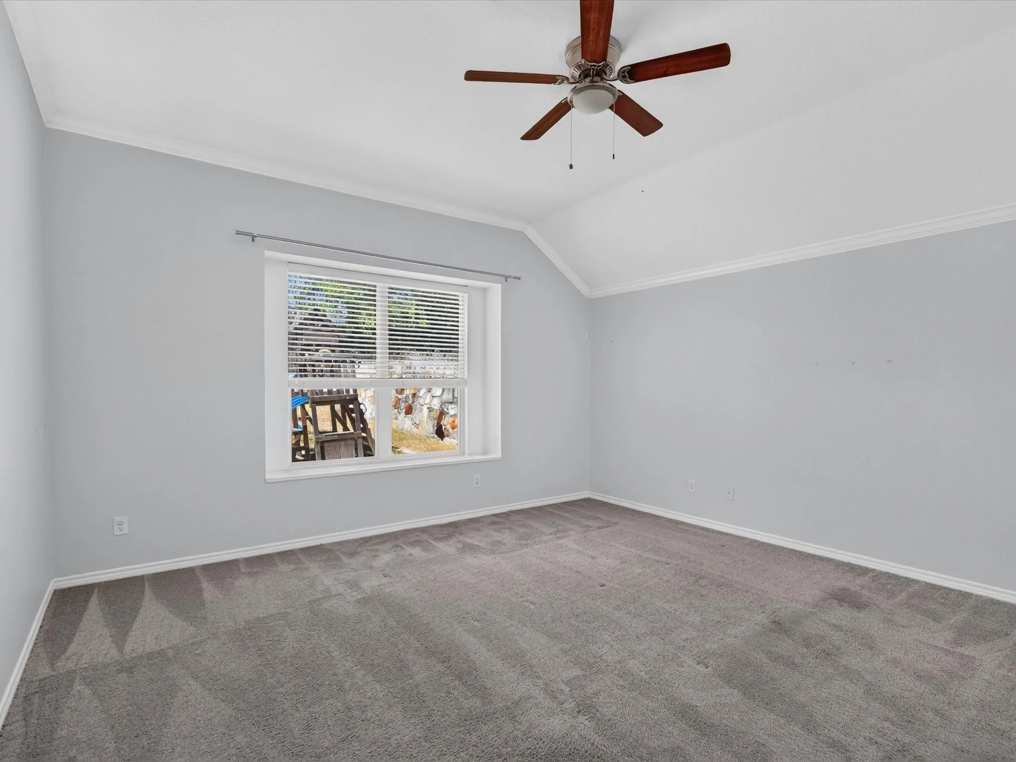 Vaulted ceiling with crown molding, ceiling fan with wood-finish blades, window with blinds, and gray carpeting