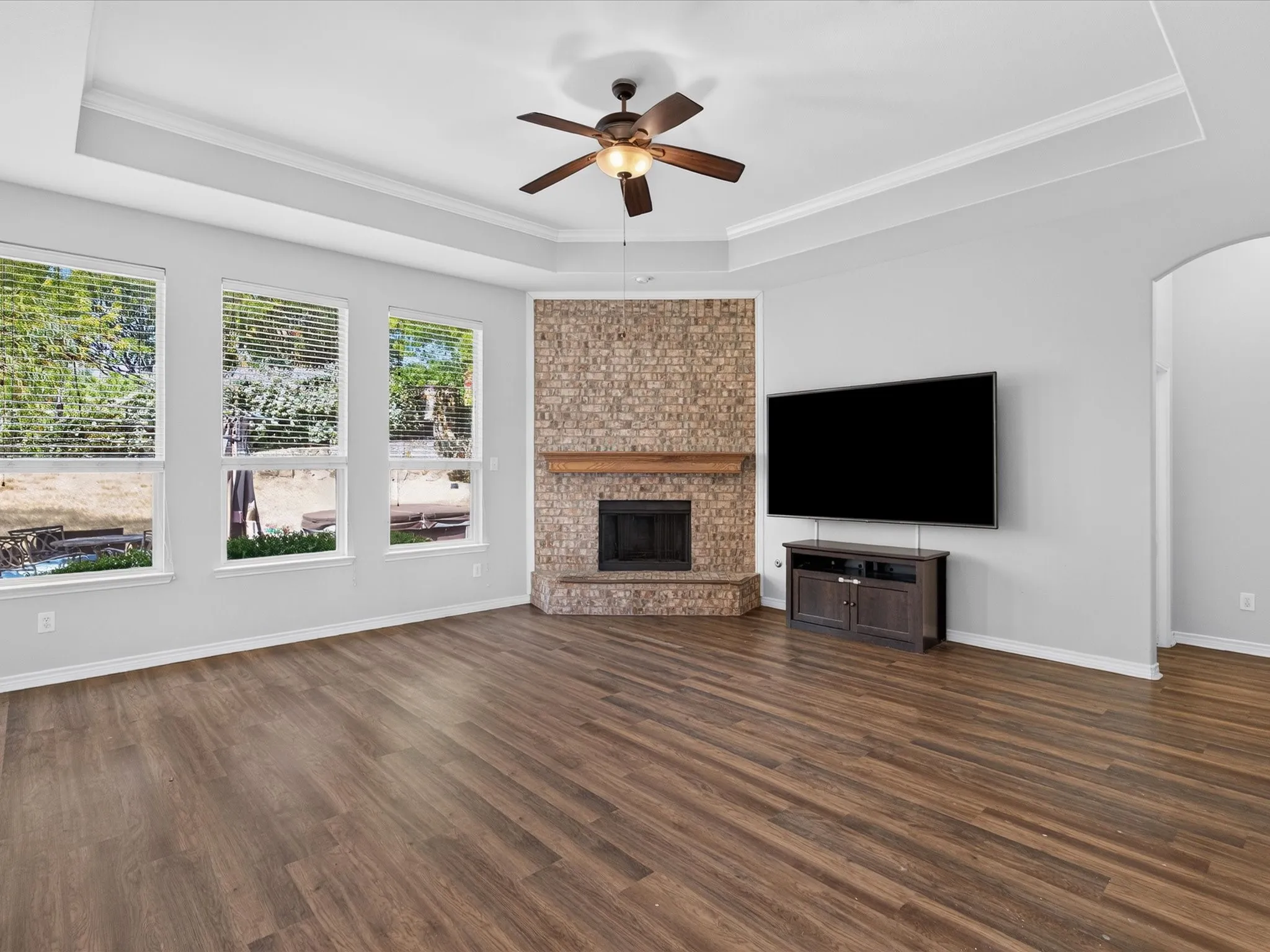 Wood-finish flooring, tray ceiling with crown molding, ceiling fan, corner brick fireplace with wood mantel, and multiple windows