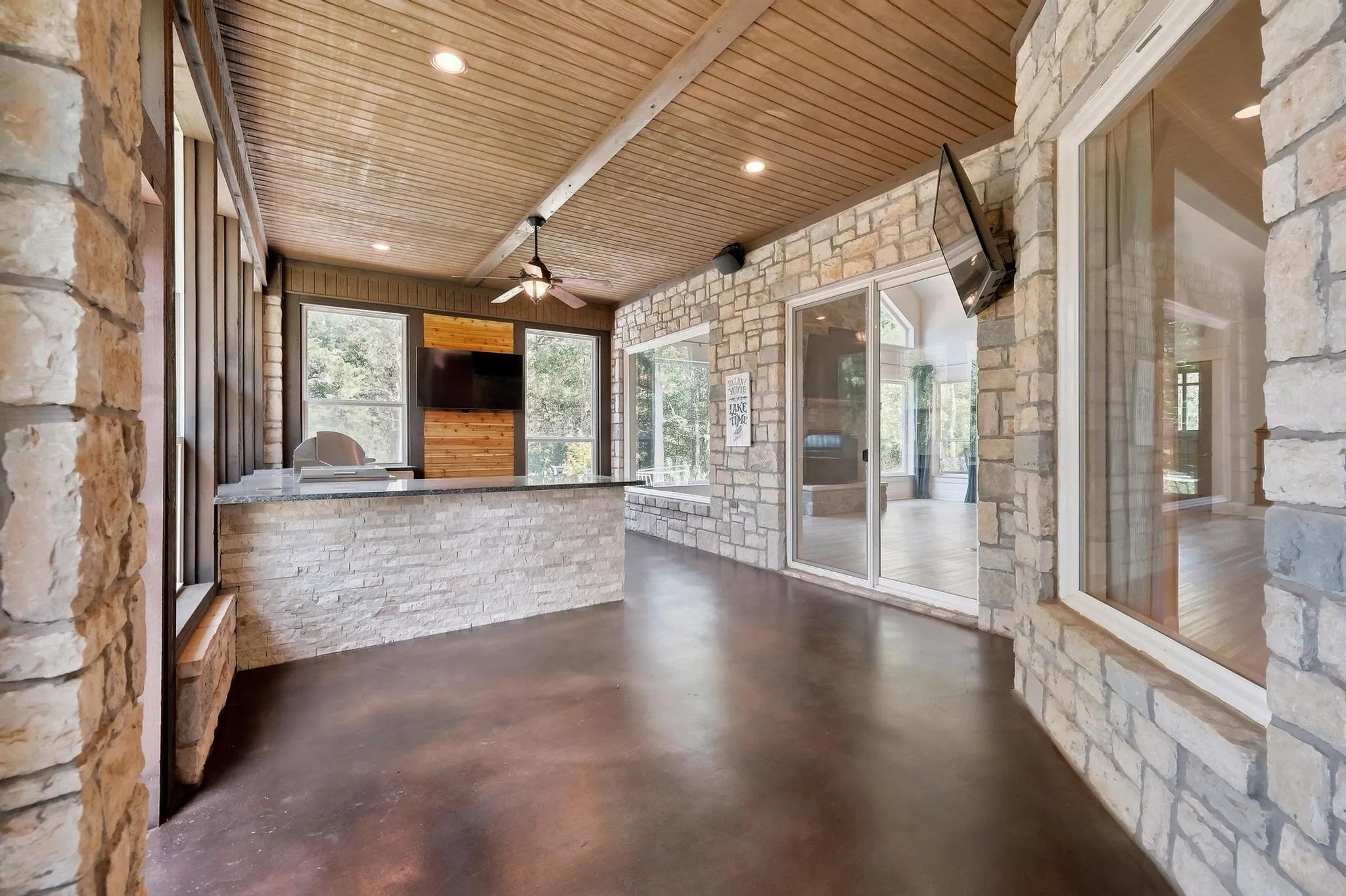 Stone veneer walls with large window openings, wood plank ceiling, and a built-in outdoor kitchen area featuring a grill and counter space