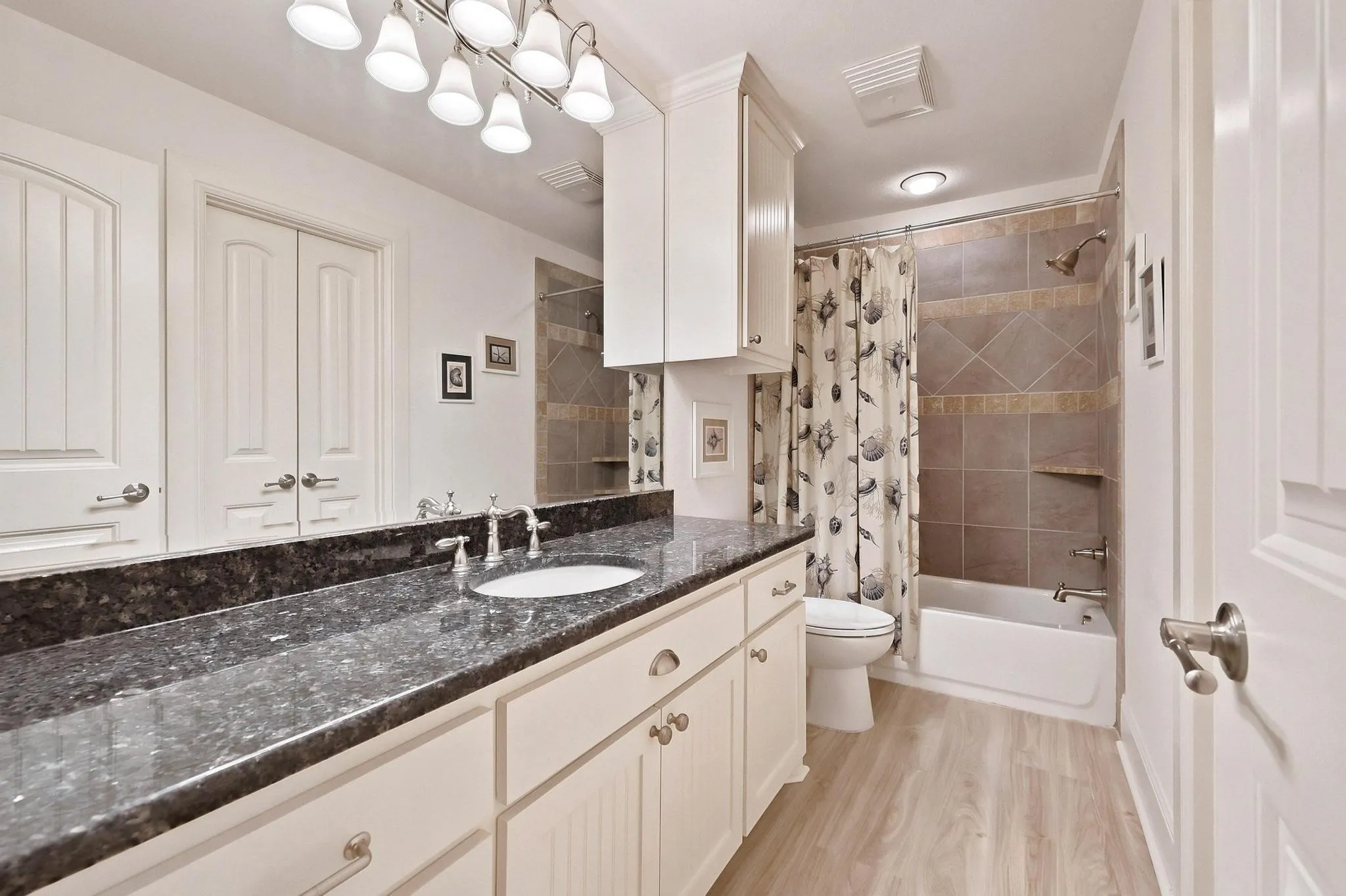 Vanity with a single undermount sink and dark countertop, white shaker-style cabinetry, wood-finish flooring, and a tiled shower-tub combination