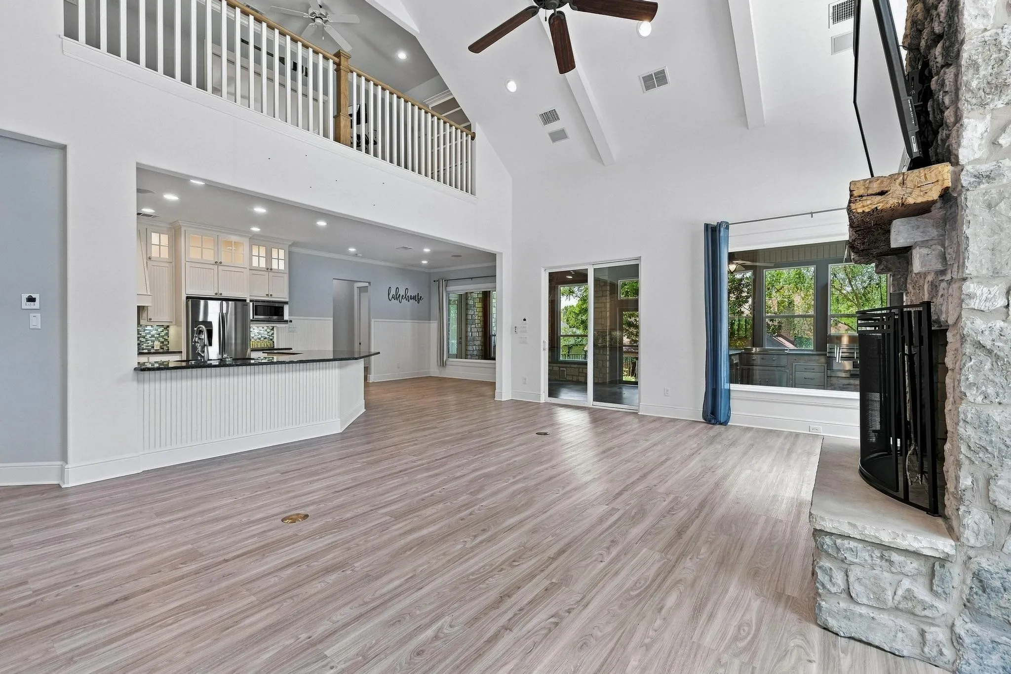 Vaulted ceiling with exposed beams, wood-finish flooring, stone fireplace with a wood mantle, kitchen island with dark countertops, and a second-story balcony overlooking the space
