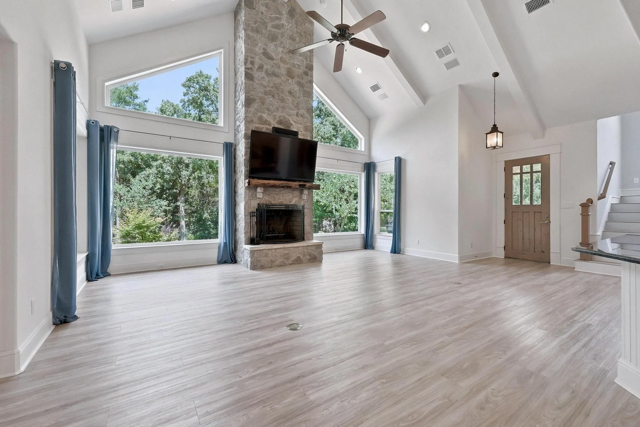 Vaulted ceiling with exposed beams, stone fireplace, wood-finish flooring, large picture windows, and a craftsman-style entry door