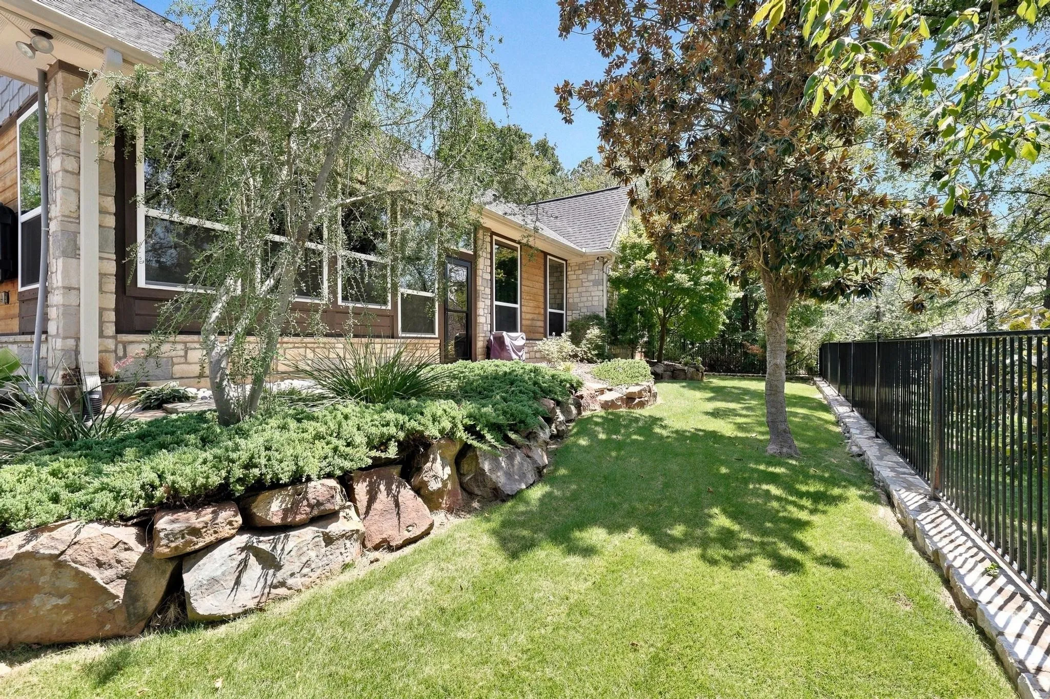 Stone and siding exterior with multiple large windows, rock retaining wall, mature landscaping, and a black wrought iron fence