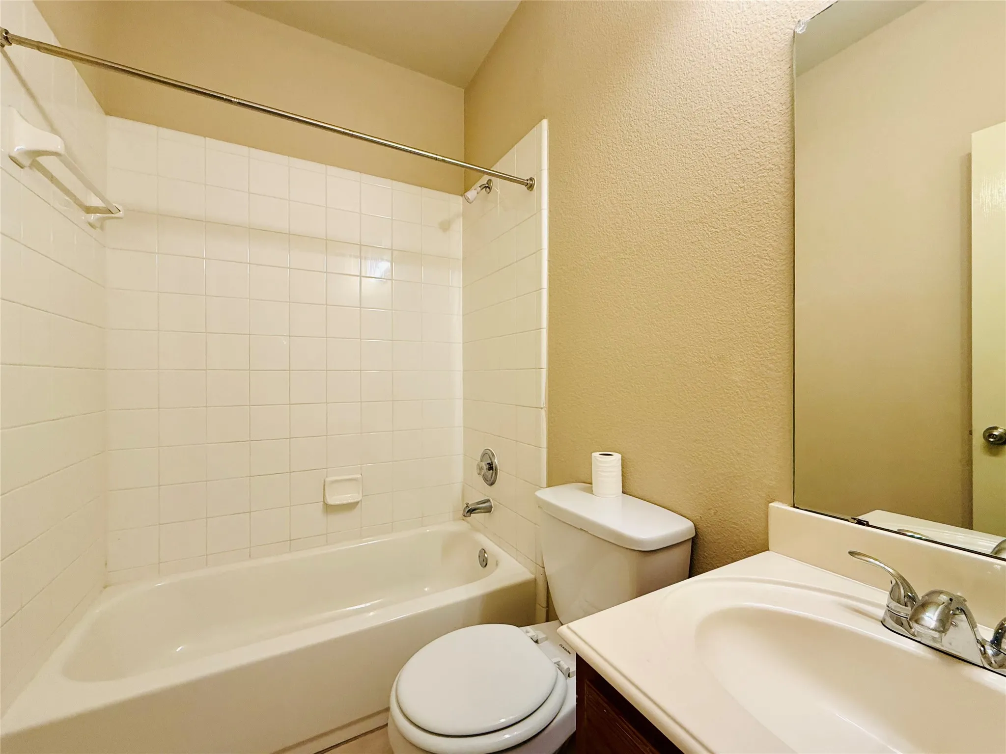 Built-in bathtub with white tile surround, chrome shower rod, wall-mounted mirror, and vanity with integrated sink