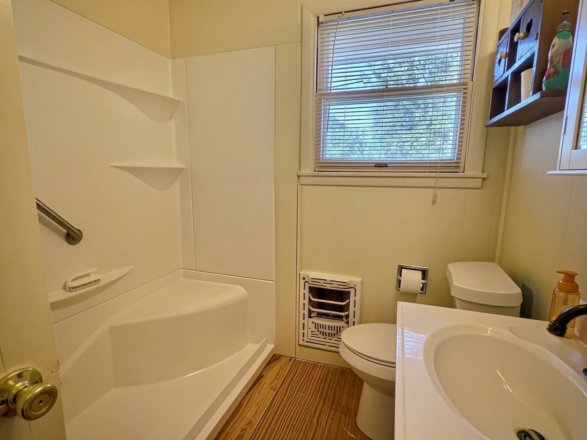 Integrated shower stall with built-in shelving, wood-finish flooring, white vanity with integrated sink, and window with privacy blinds