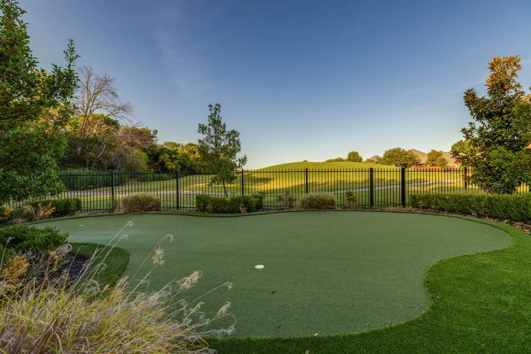 Artificial turf putting green with two cups, bordered by manicured shrubs and a black metal fence, overlooking a landscaped berm