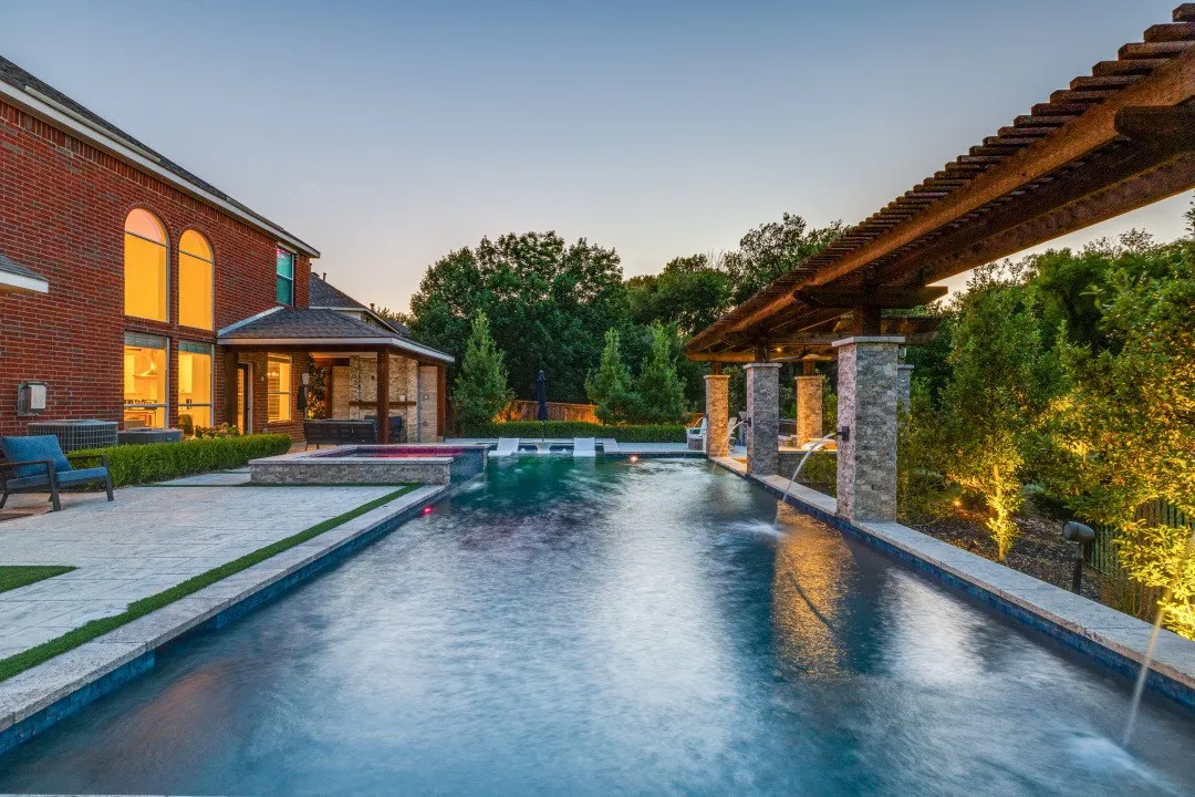 Rectangular swimming pool with integrated spa and water features, surrounded by extensive hardscaping and a covered gazebo