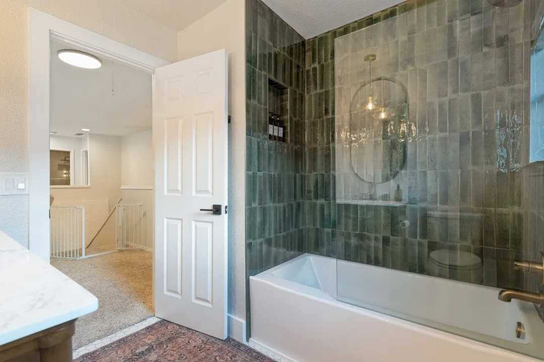 Bathtub with a glass partition, green subway tile surround, recessed shelf, and brass-finish fixtures