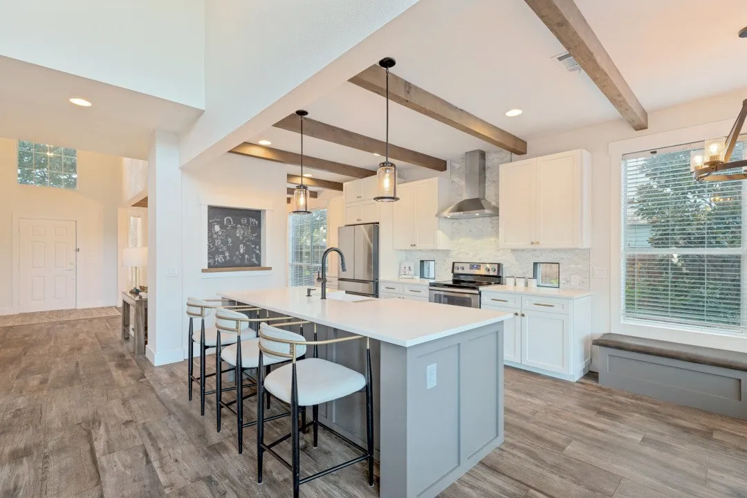 Open-concept kitchen featuring an expansive central island with seating, wood-finish flooring, white cabinetry, stainless steel appliances, and decorative ceiling beams