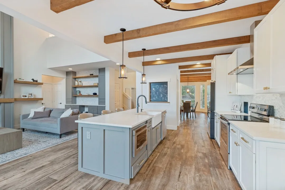 Exposed ceiling beams, wood-finish flooring, white cabinetry, a kitchen island with built-in appliances, and a farmhouse sink