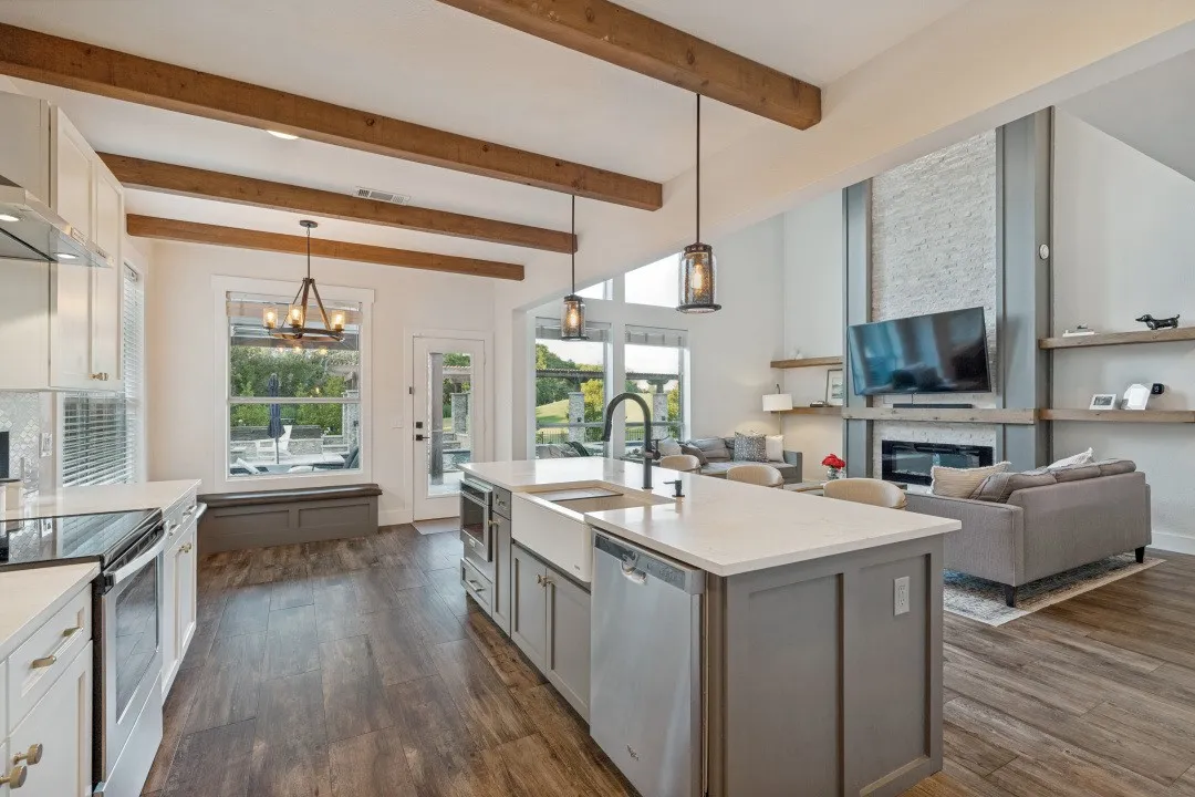 Open-concept layout with exposed wood beams, wood-finish flooring, a kitchen island with a farmhouse sink, and a modern fireplace with a stone facade
