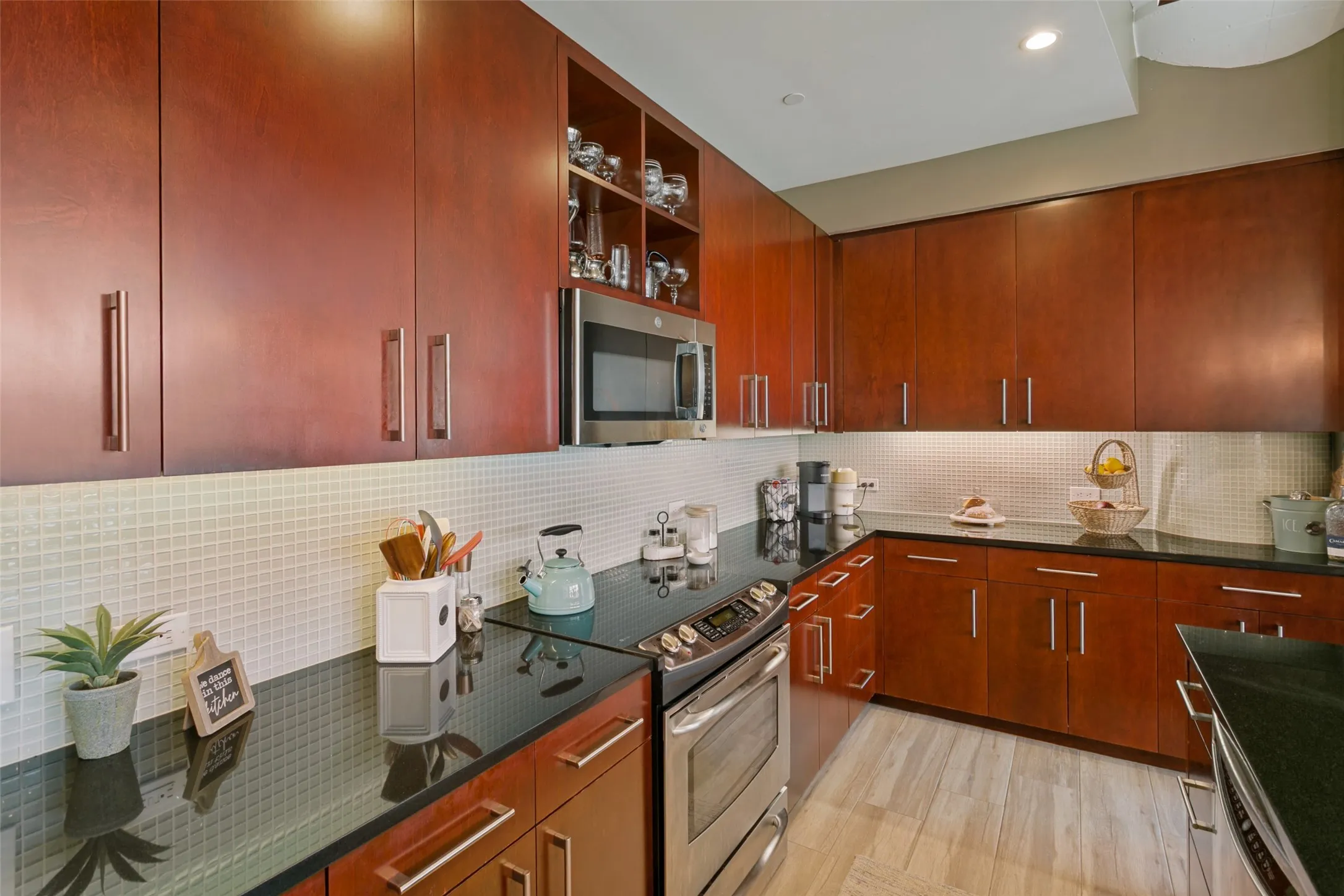 Cherry wood-finish cabinetry with brushed nickel hardware, black solid surface countertops, stainless steel appliances, and mosaic tile backsplash