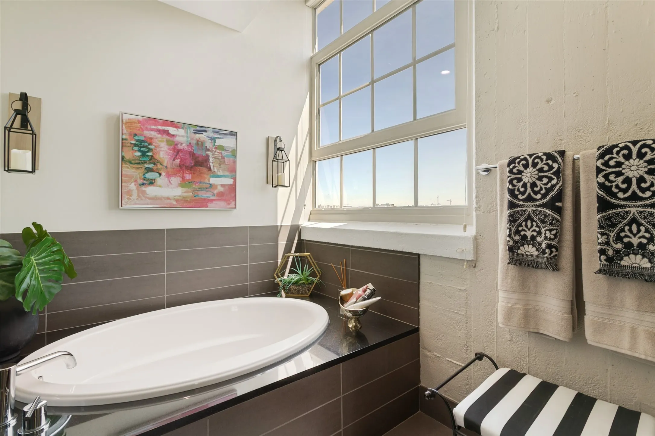 Deep soaking tub, dark tile surround, large multi-pane window, wall-mounted sconce lighting, and towel bar