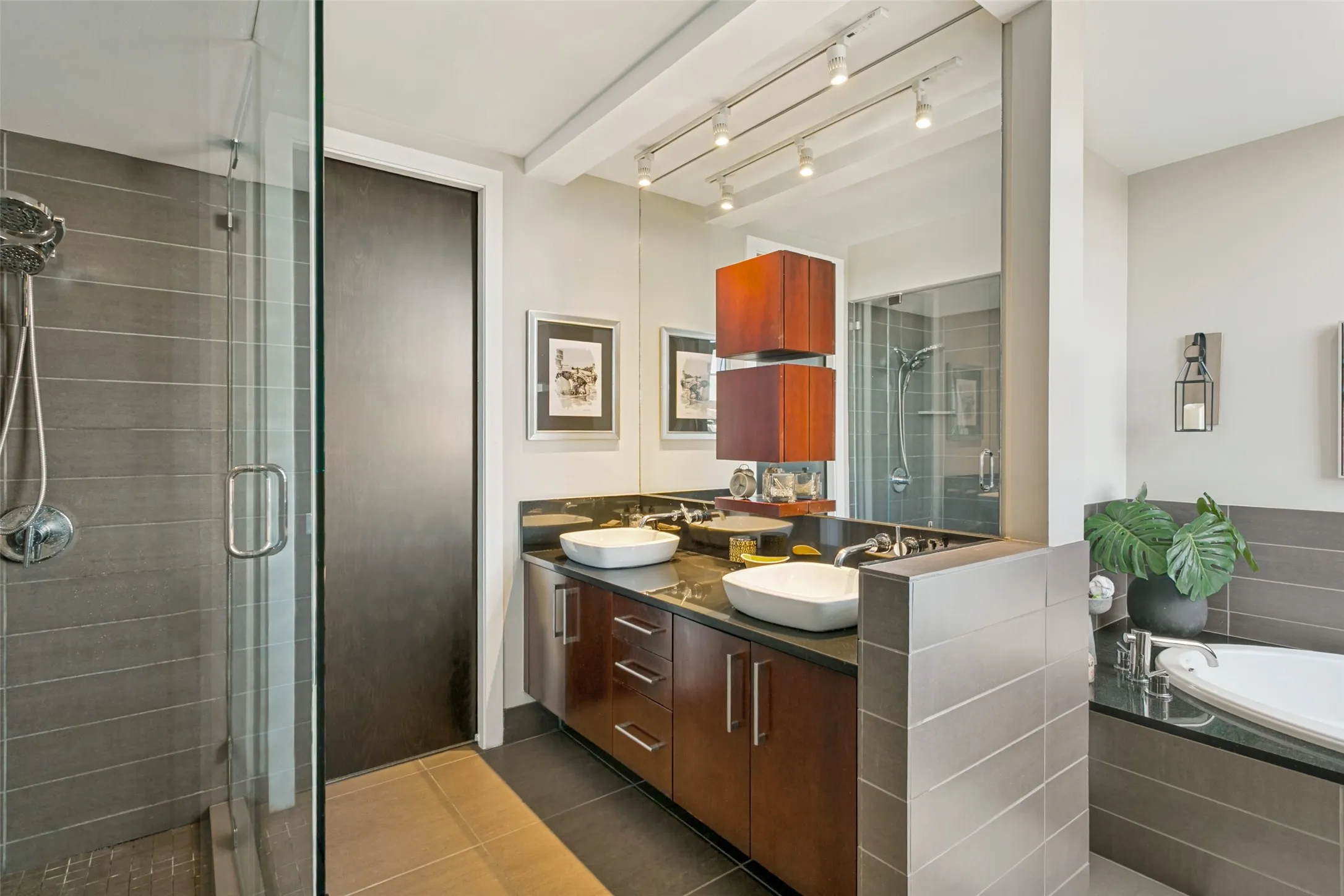 Glass-enclosed shower with subway tile surround, dual vanity with vessel sinks, dark wood-finish cabinetry, and track lighting
