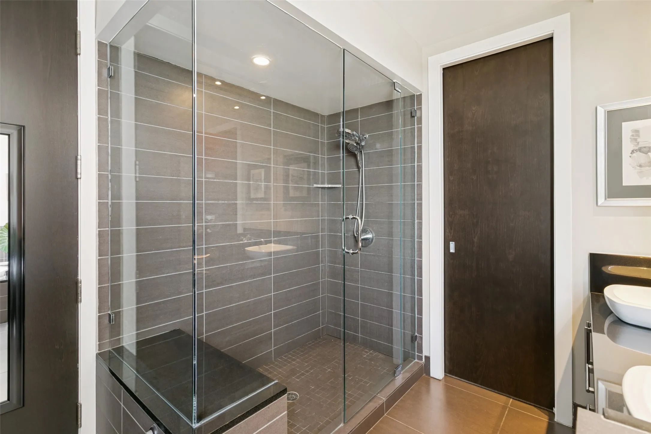 Frameless glass shower enclosure with a built-in bench, gray horizontal tile, and a handheld shower fixture