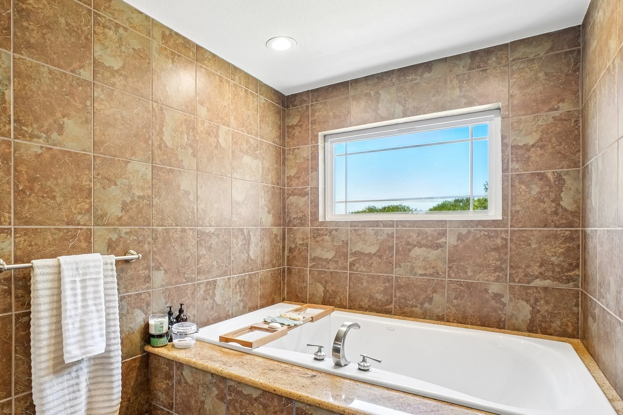 Spa-inspired primary bath featuring travertine tile surround, soaking tub with wood-finish deck, chrome faucetry, heated towel rack, recessed lighting, and a transom window framing natural light and scenic vistas